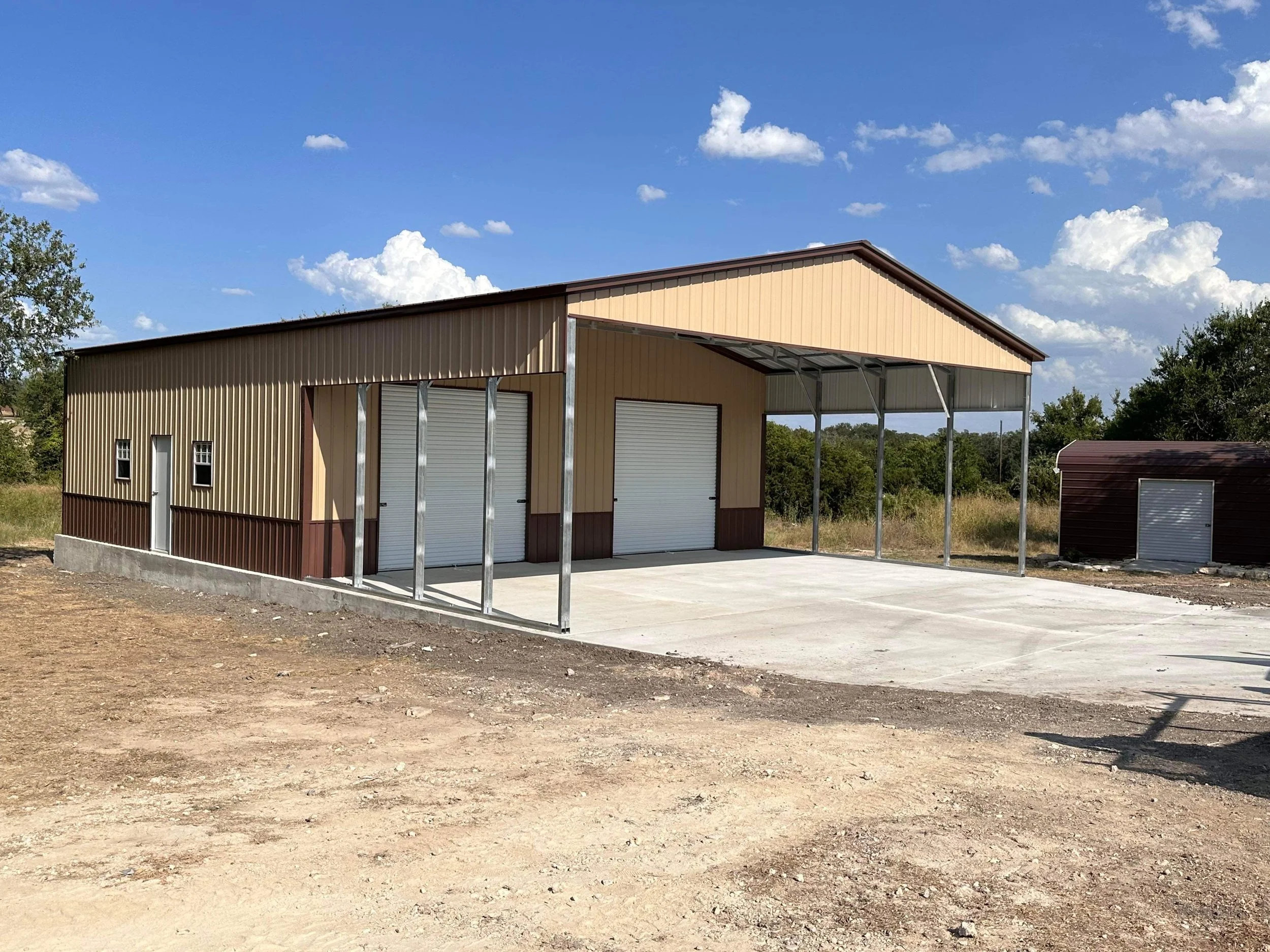 A metal building with a sloped roof and multiple roll-up garage doors, situated on a concrete pad, surrounded by grassy and dirt areas under a blue sky with scattered clouds.