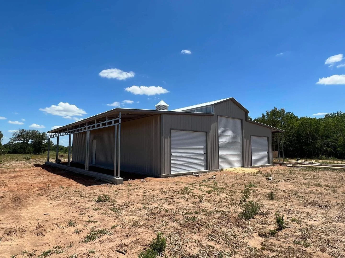 A metal building with roll-up garage doors on a dirt lot under a blue sky with clouds, surrounded by sparse vegetation and trees in the distance.