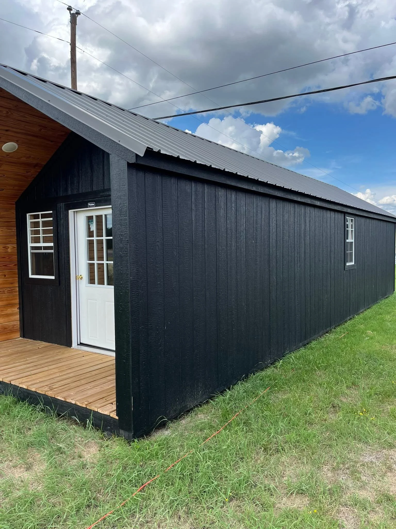 A house with black exterior walls, white window and door, and a metal roof, located on a wooden deck, with green grass and cloudy sky in the background.