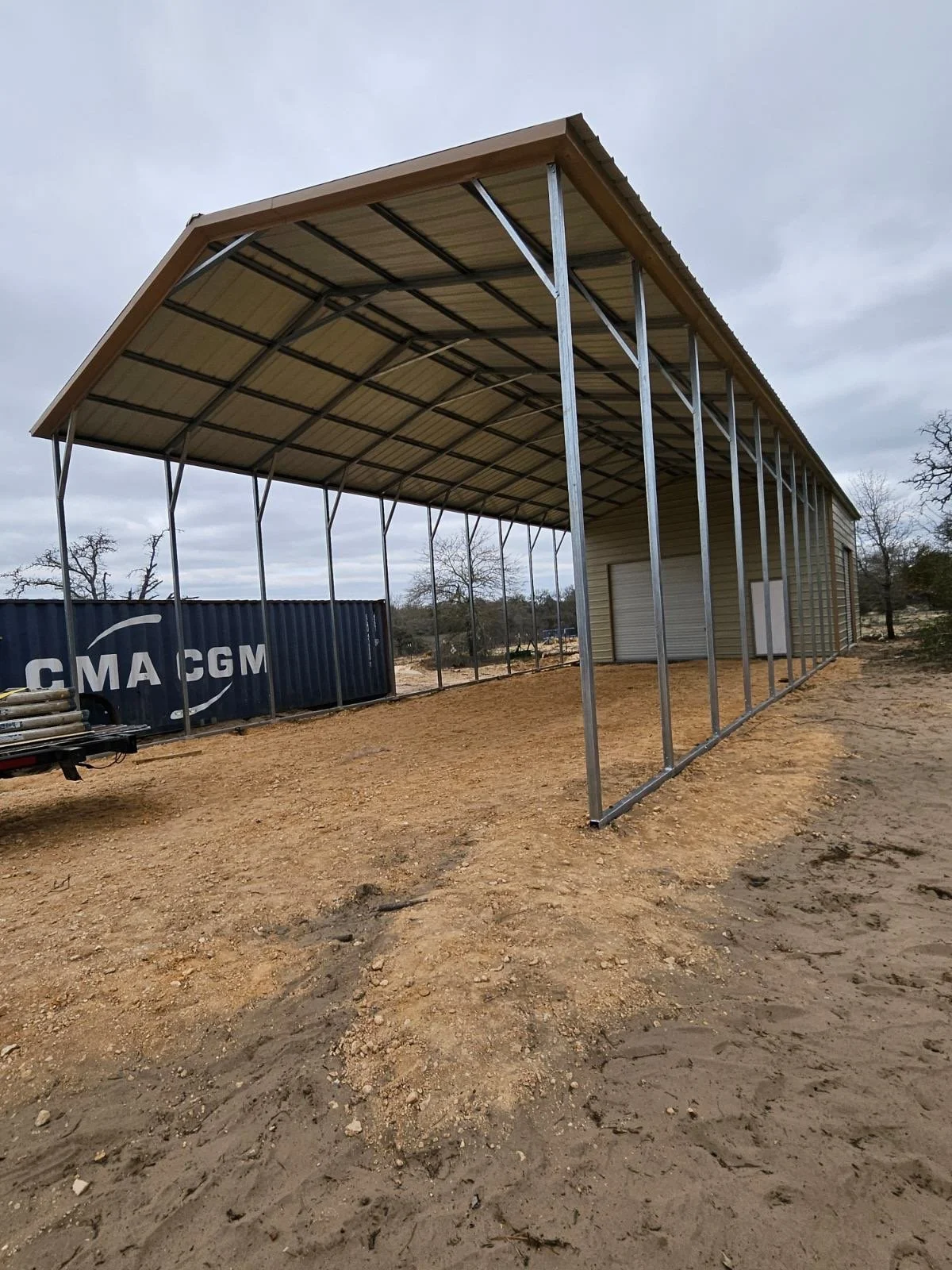 Under construction metal building with a sloped roof, metal framing, and a tan exterior, situated on a dirt lot with a shipping container reading 'CMA CGM' nearby, and cloudy sky overhead.