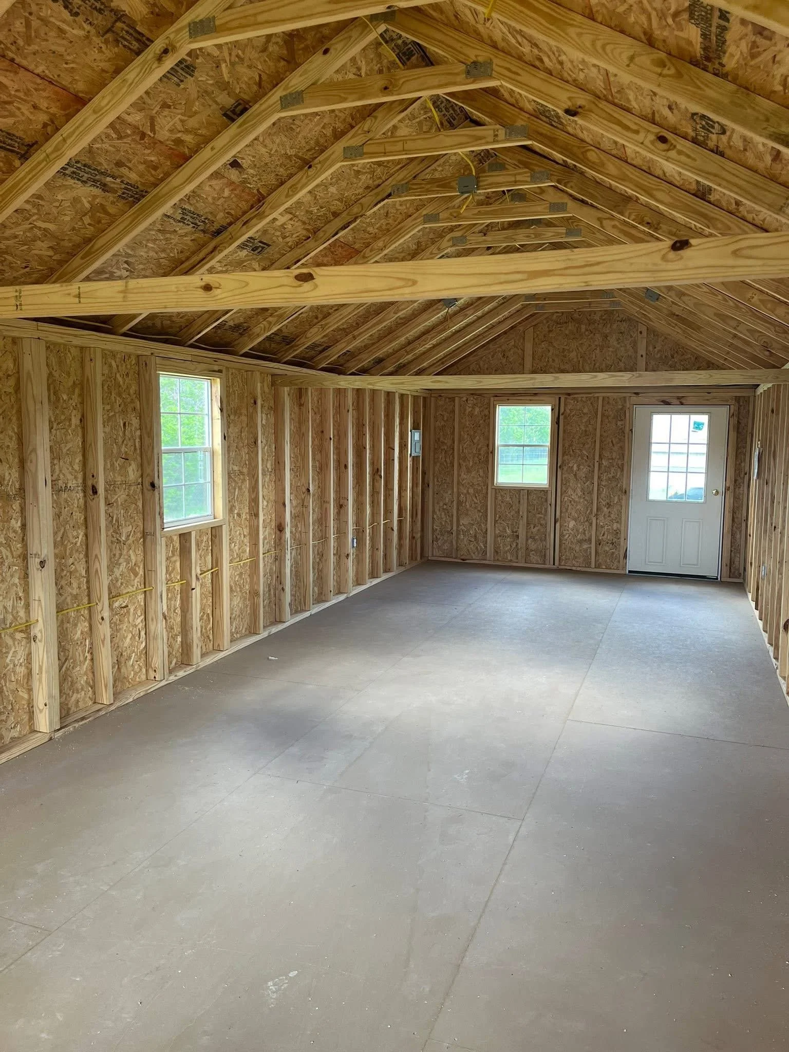 Interior of a house under construction with exposed wooden framing, plywood sheathing, and bare concrete floor, with three windows and a door.