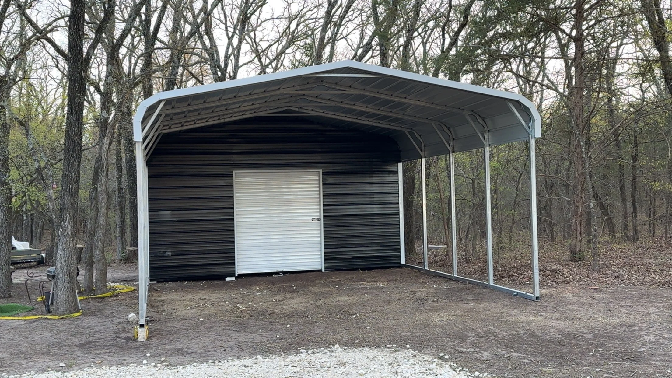 A metal storage shed with a white roll-up door, situated in a wooded area surrounded by trees with no leaves.