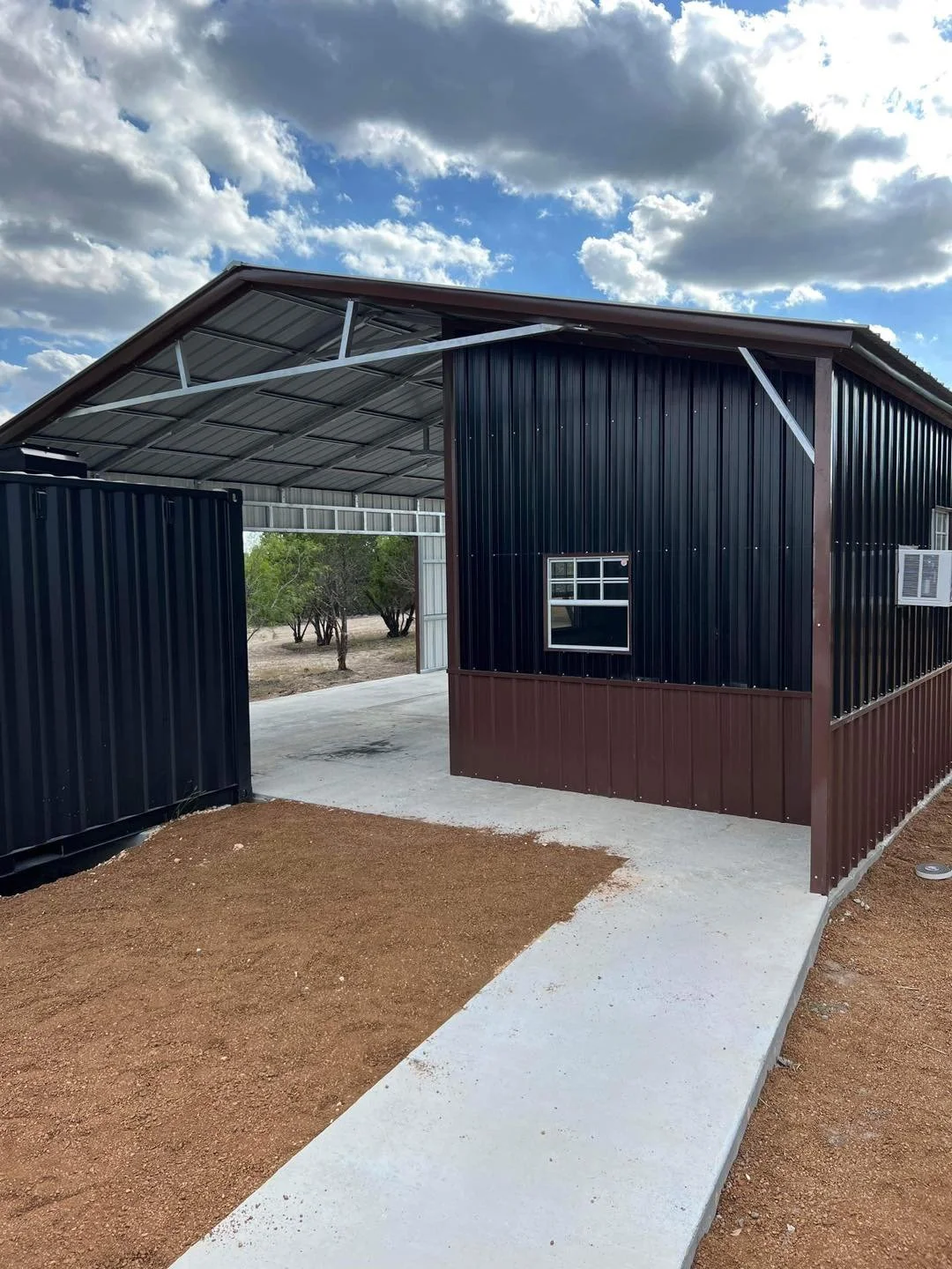 A black and brown metal shed with a small window, a ramp, and a covered area under a cloudy sky.