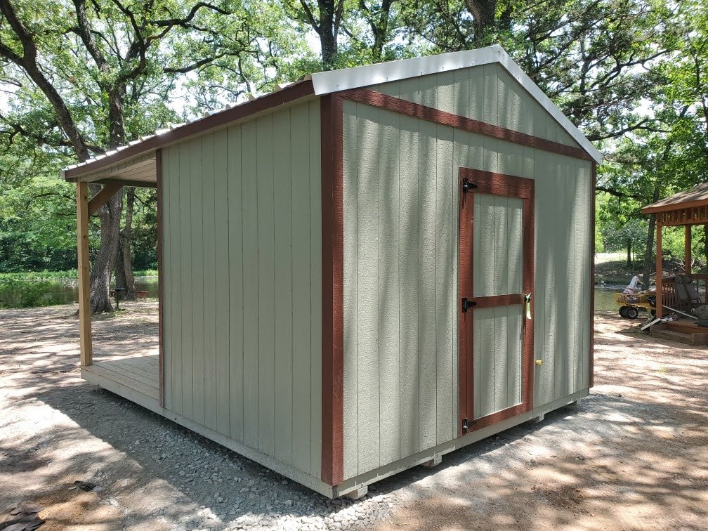 A small shed with beige walls and brown trim, featuring a door with a window, set on a gravel surface in a wooded area.