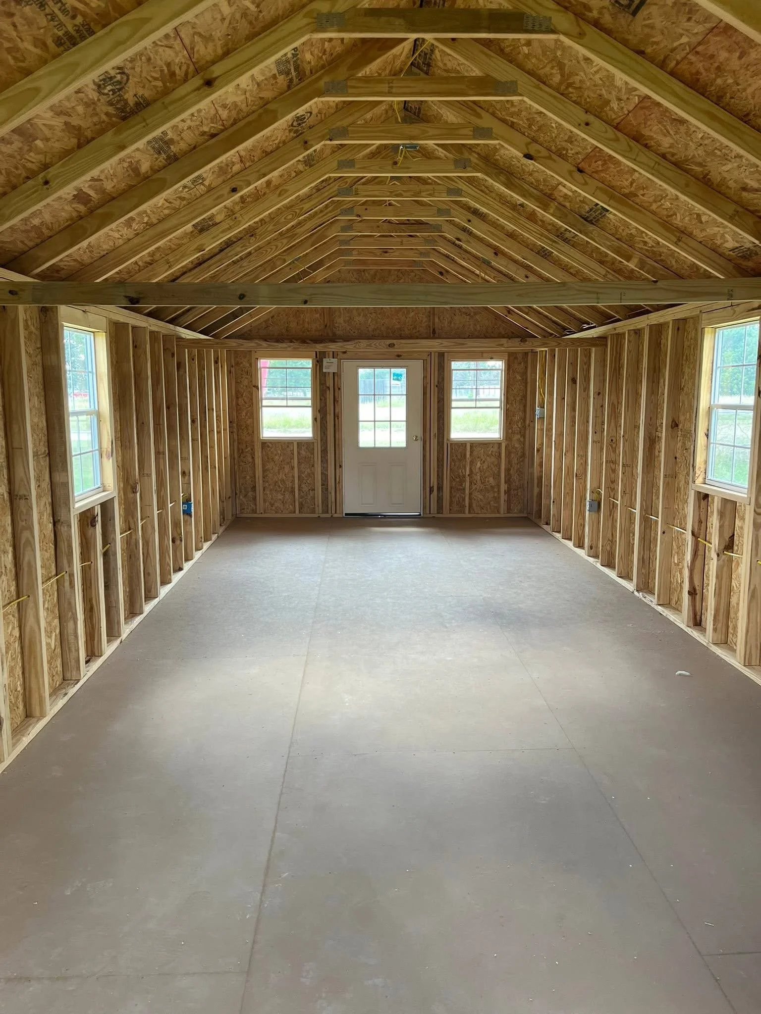 Interior of a building under construction with exposed wooden framing, two windows, and a door at the far end. The roof has wooden rafters and a basic electrical wiring setup visible.