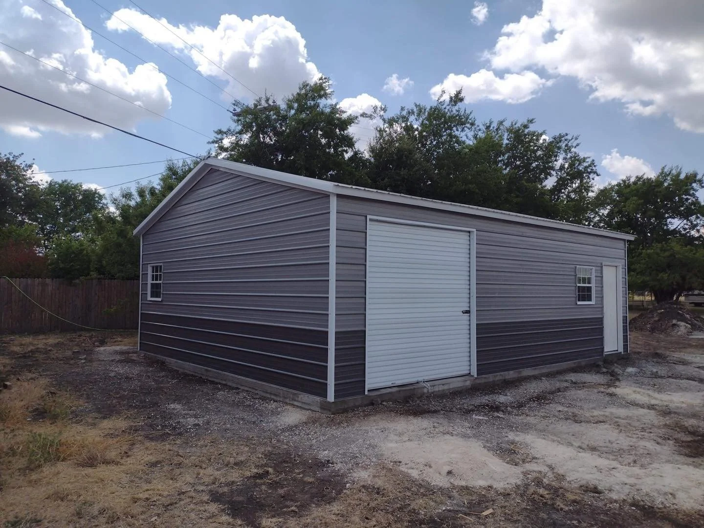 A gray metal storage shed with a white roll-up door and a white pedestrian door, situated on a patch of dirt and gravel, surrounded by trees and a wooden fence, under a blue sky with scattered clouds.