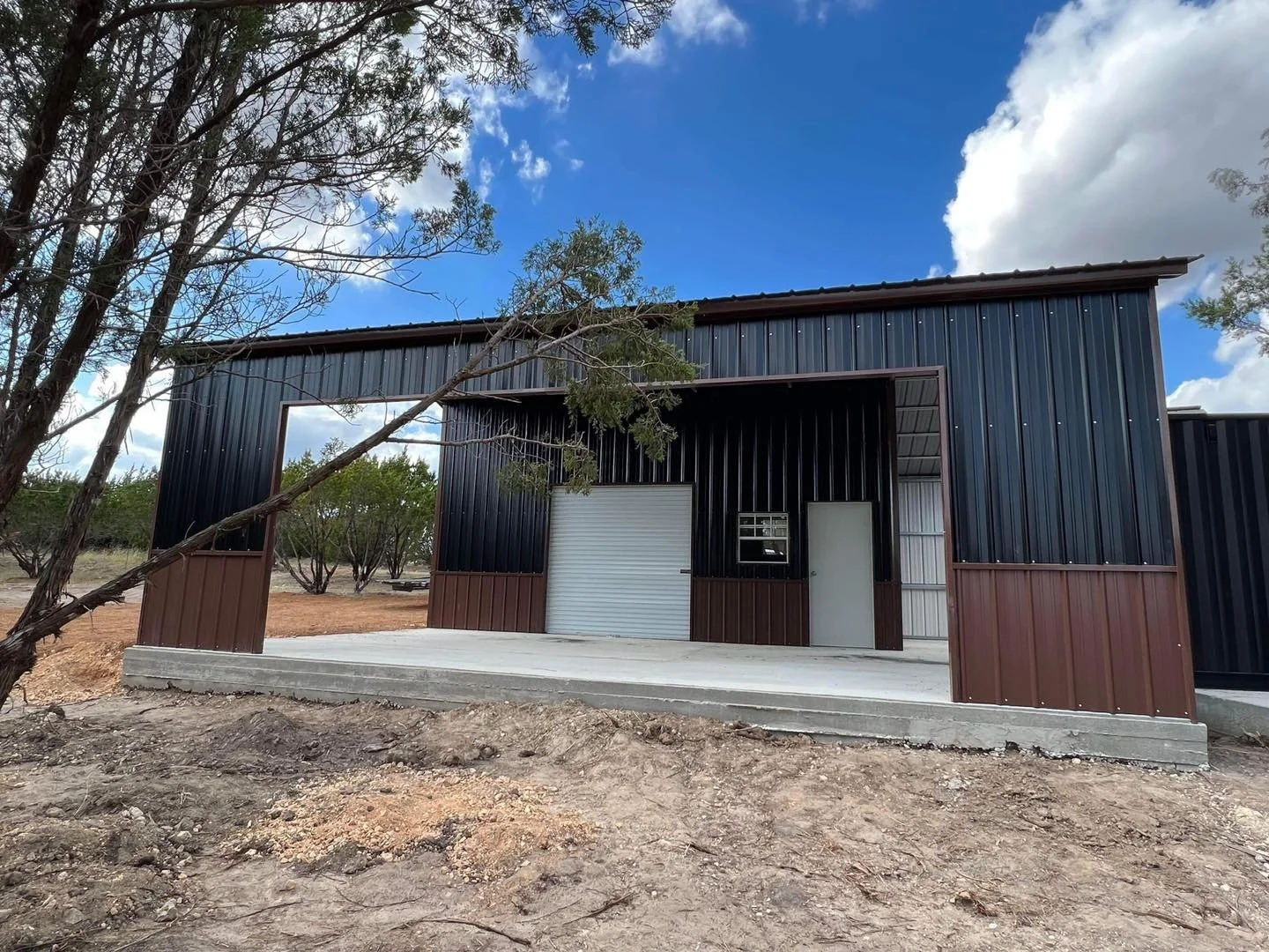 A metal building with a black and brown exterior, a large roll-up garage door, a smaller side door, and a small window, located in a rural area with trees and dirt ground.