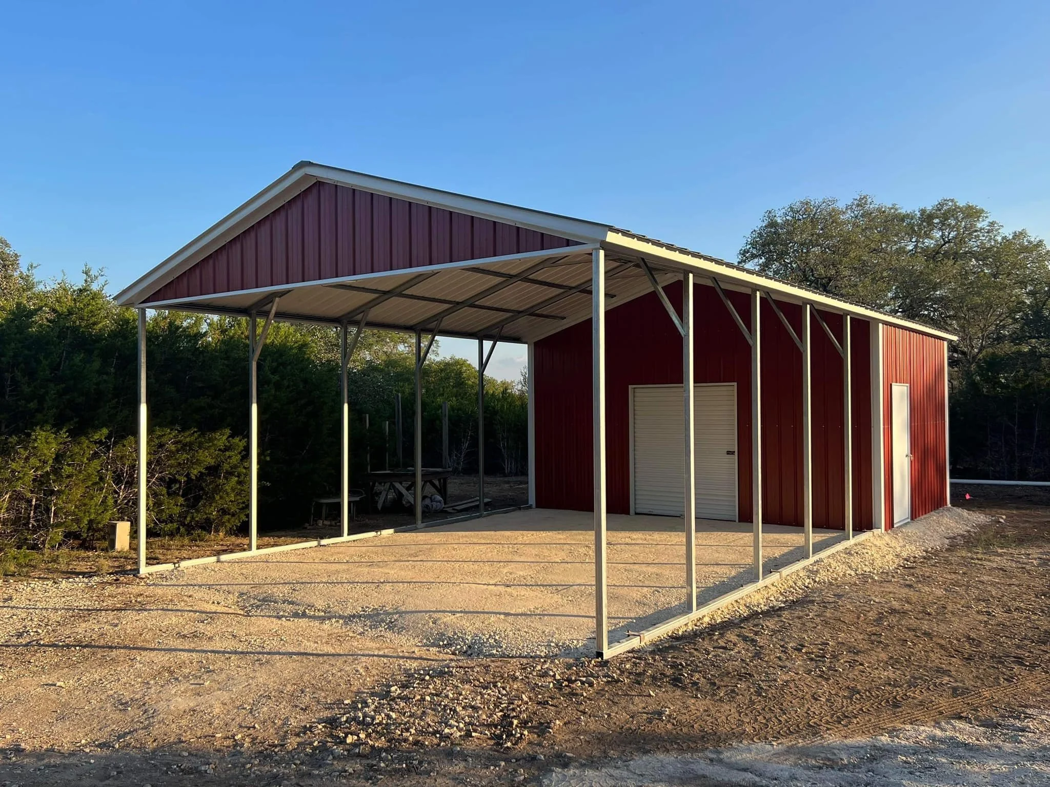 A red metal building under construction with a metal roof and a large door, surrounded by trees and a gravel ground, during late afternoon with clear blue sky.