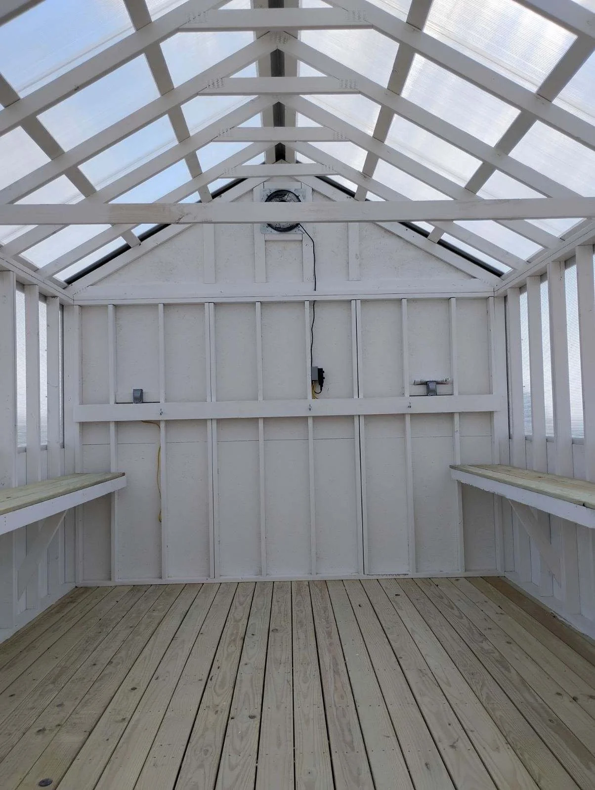 Interior of a partially completed small wooden structure with a sloped roof and built-in benches on each side. The ceiling has exposed beams and a fan at the apex. The wood is painted white, with some unpainted wooden flooring at the bottom.