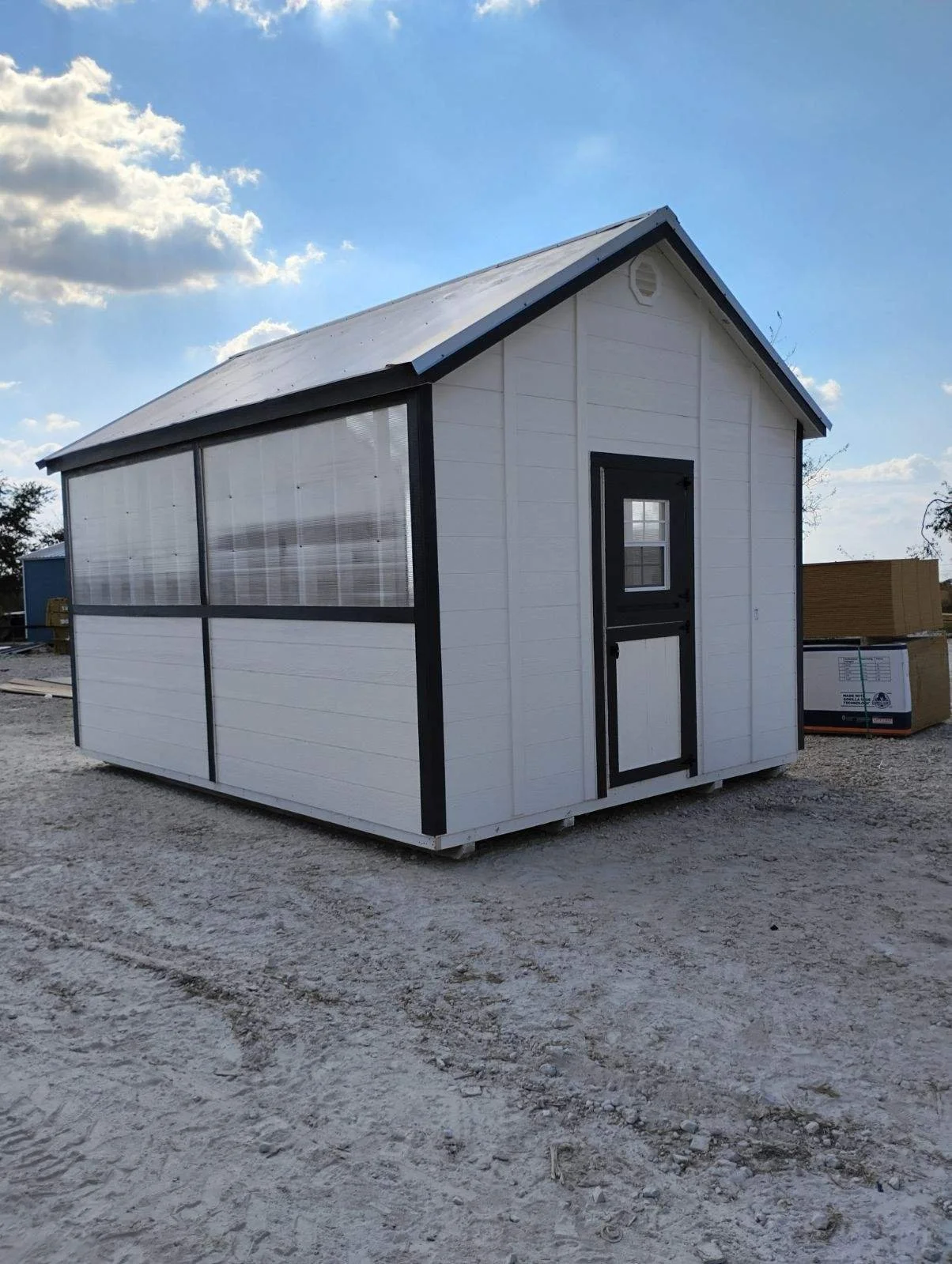 A small white farm-style shed with black trim, a small window and a black door, situated on a gravel lot on a clear day.