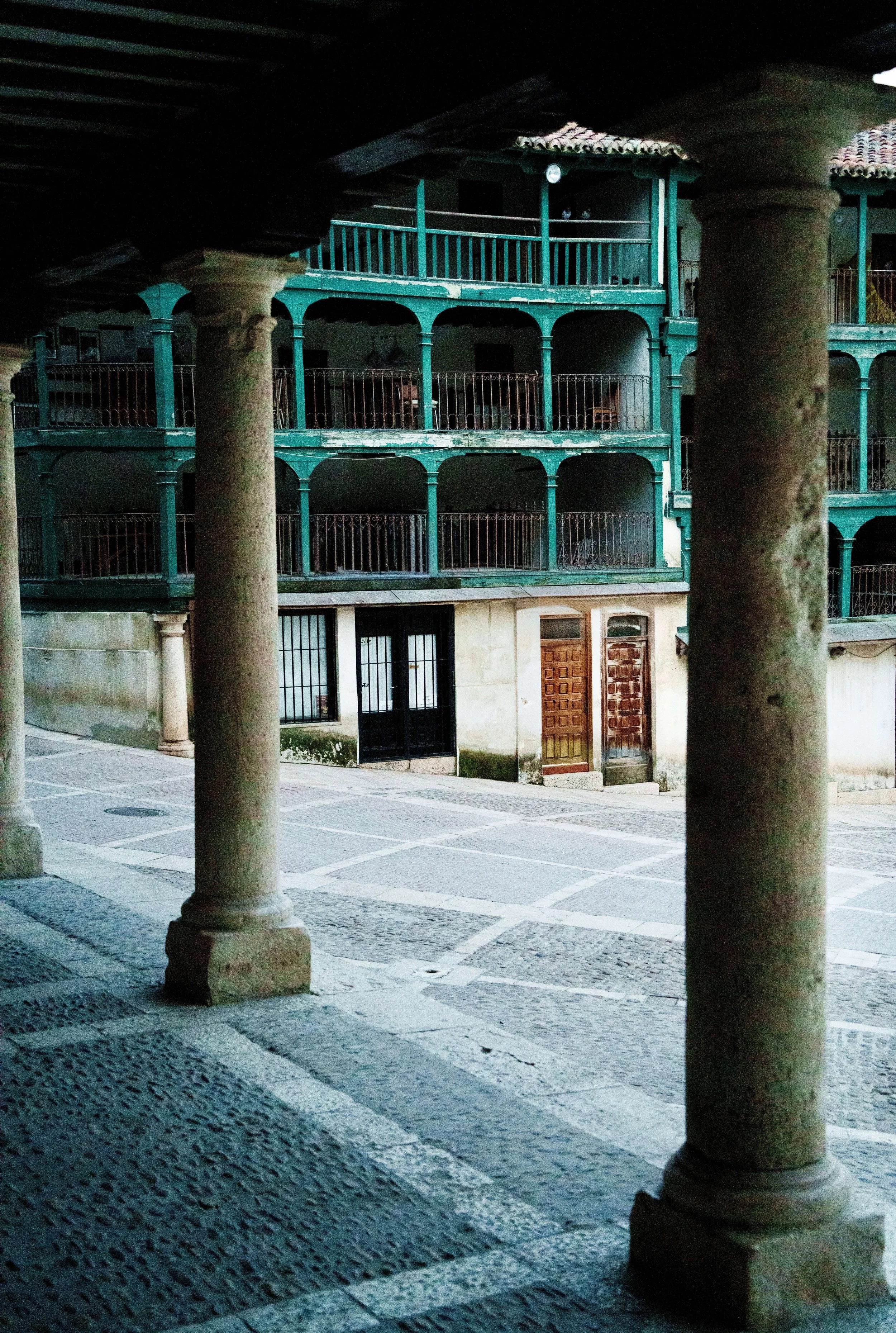 View of a multi-story building with balconies and a tiled roof, seen from behind stone columns in an outdoor courtyard at dusk or early evening.