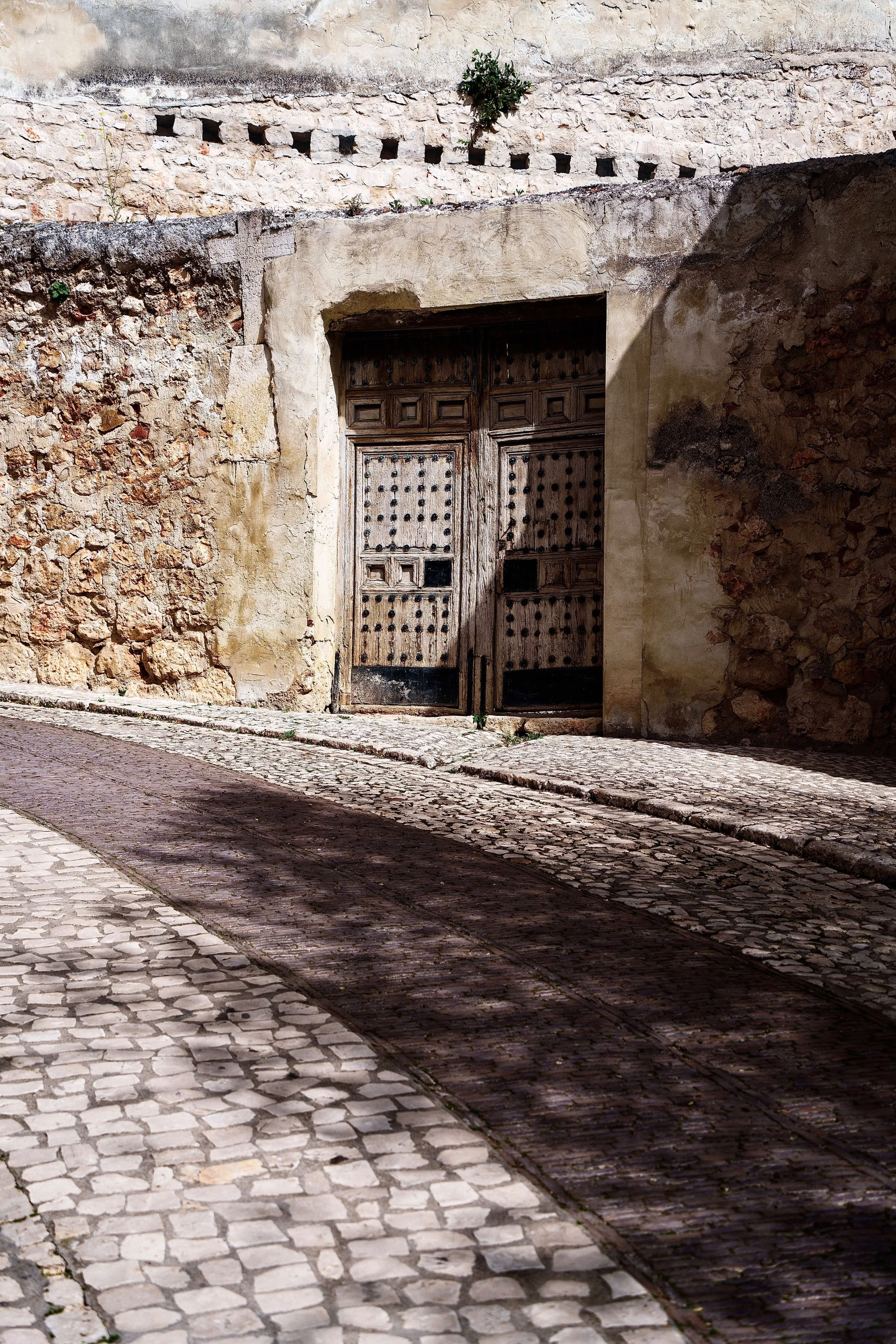 Old wooden door set into a stone and brick wall, casting a shadow on cobblestone street in daytime.