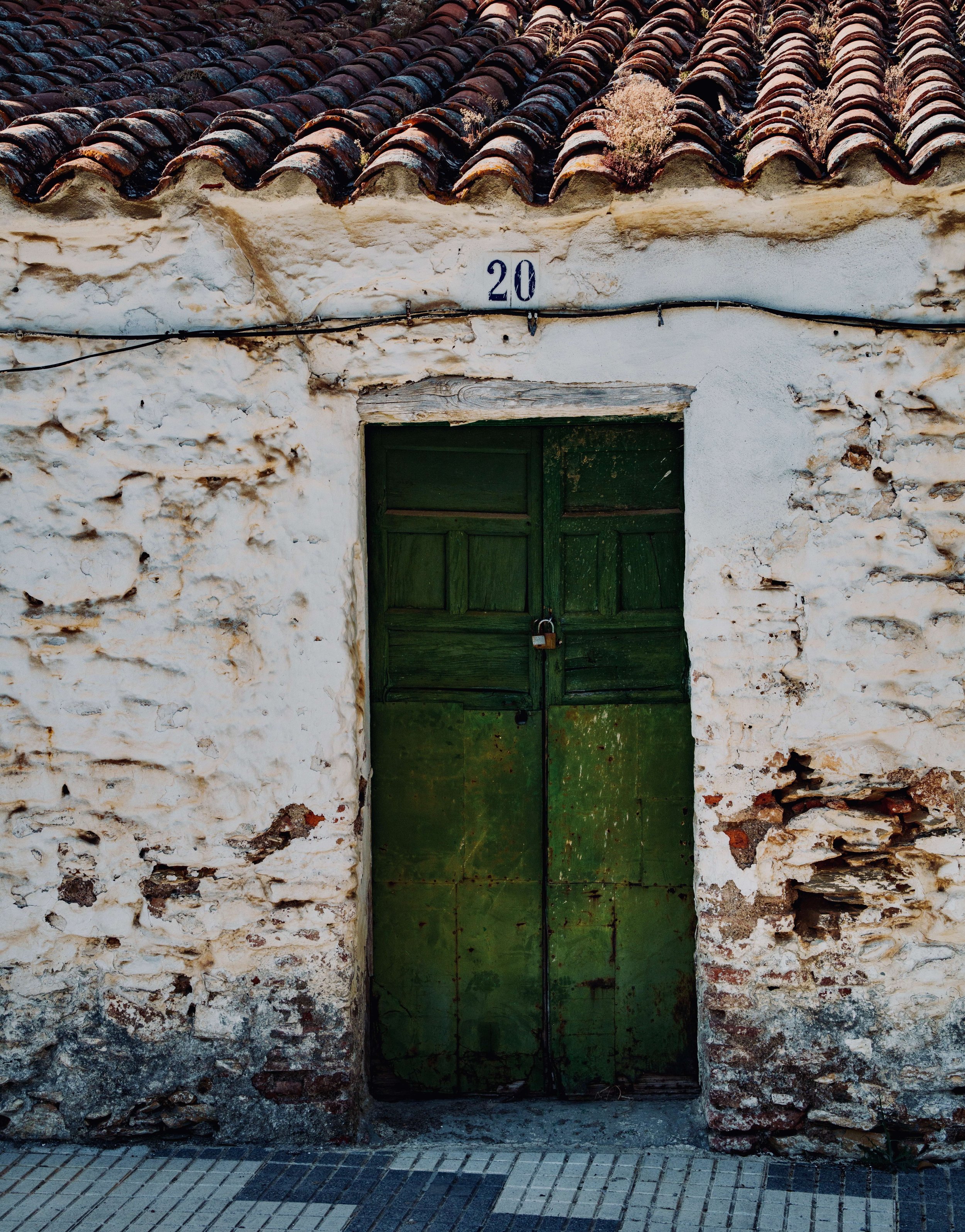 A weathered, rustic house with a terracotta tiled roof showing signs of age and moss growth, a white plastered facade with cracks and textures, a green wooden door secured with a padlock, and a stone-paved sidewalk in front.