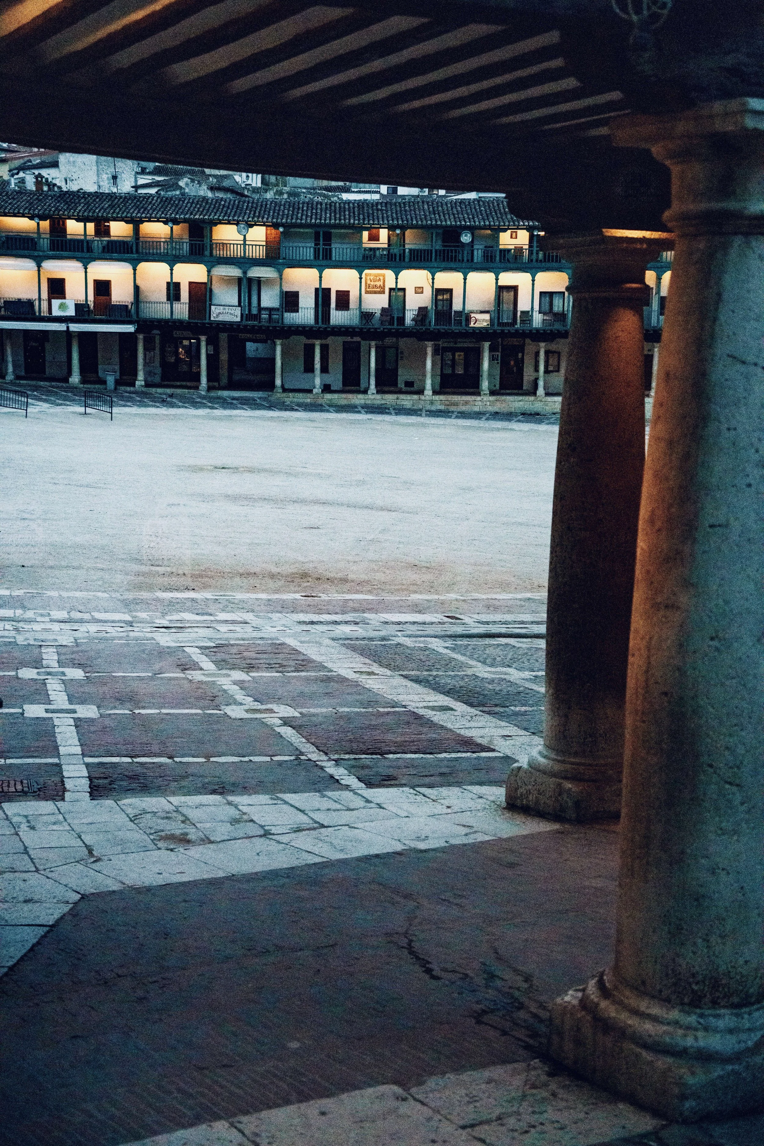 A view of a historic building with two floors, illuminated by outdoor lights, seen from under a stone archway supported by columns, with an empty square or open plaza in the foreground.