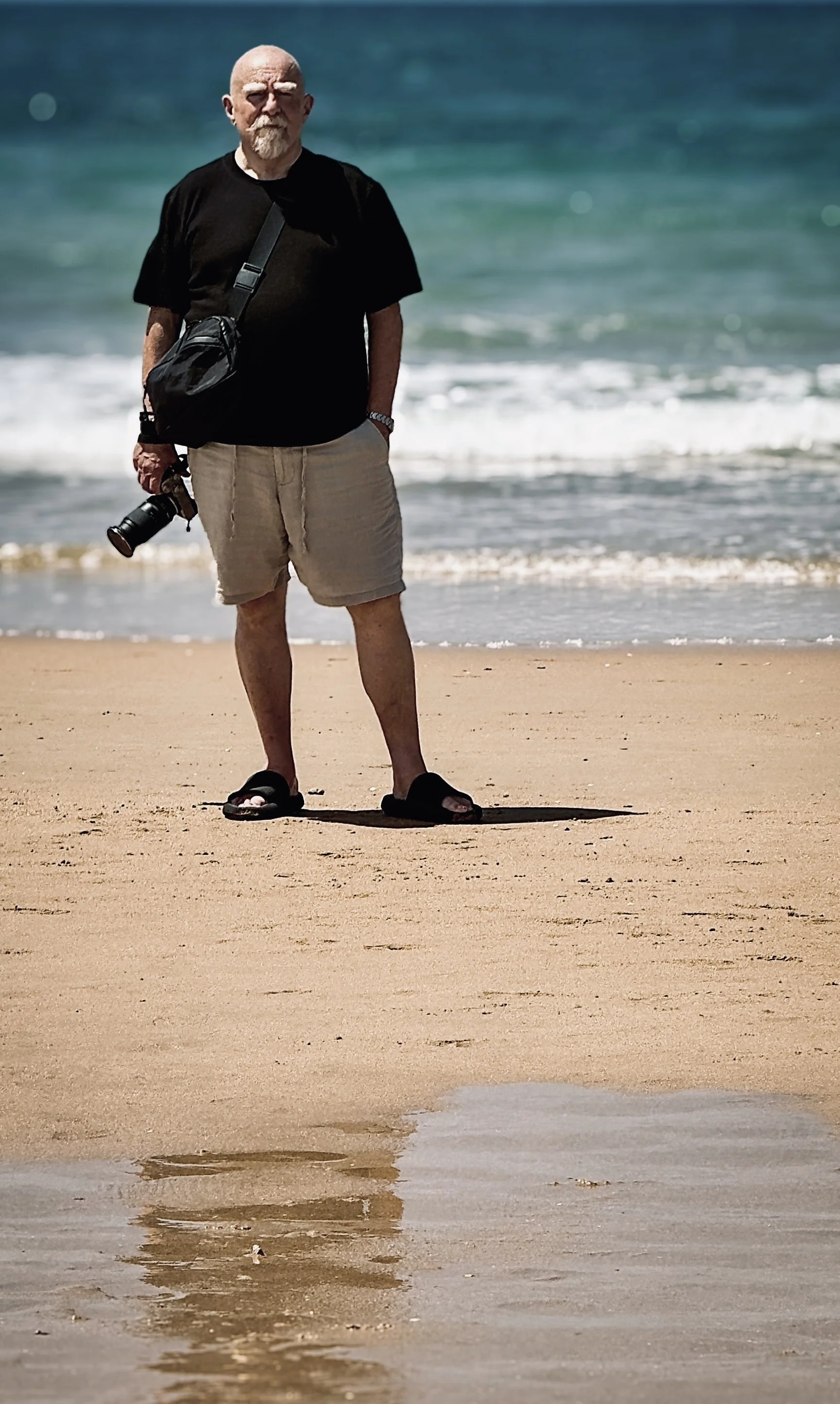 An elderly man with a beard and sunglasses standing on a sandy beach, holding a camera in his right hand, with the ocean in the background.