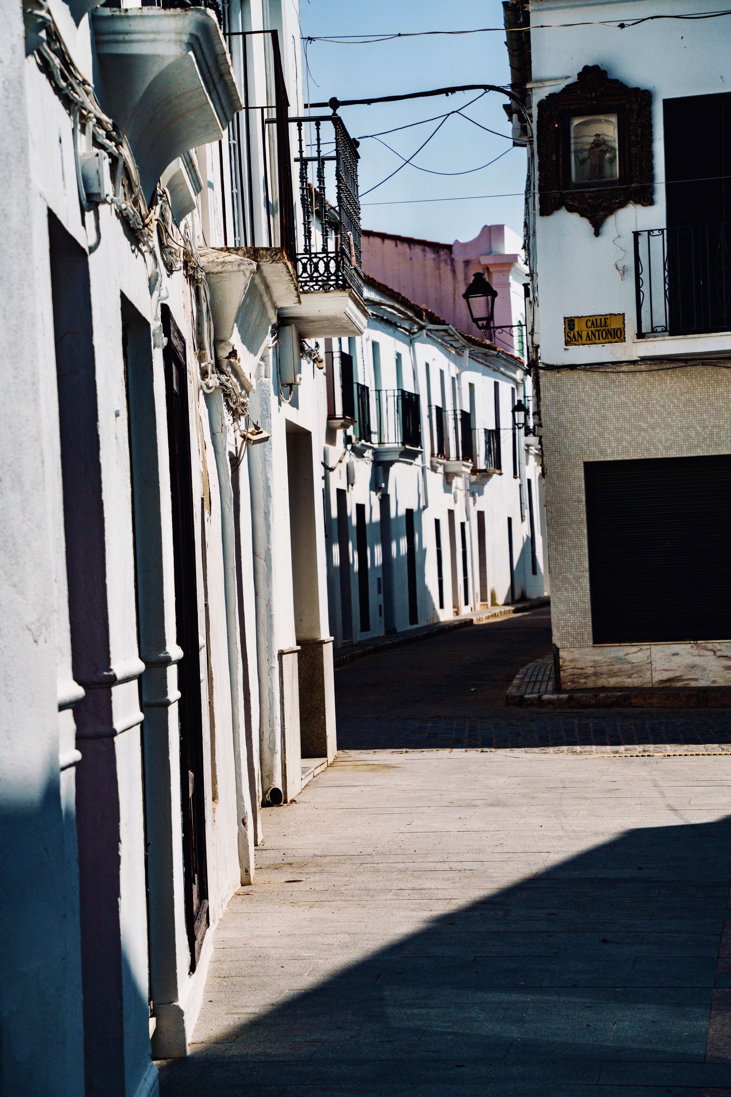 Empty street with white colonial-style buildings, black wrought iron balconies, and a street sign reading 'Calle San Antonio', with sunlight casting shadows on the sidewalk.