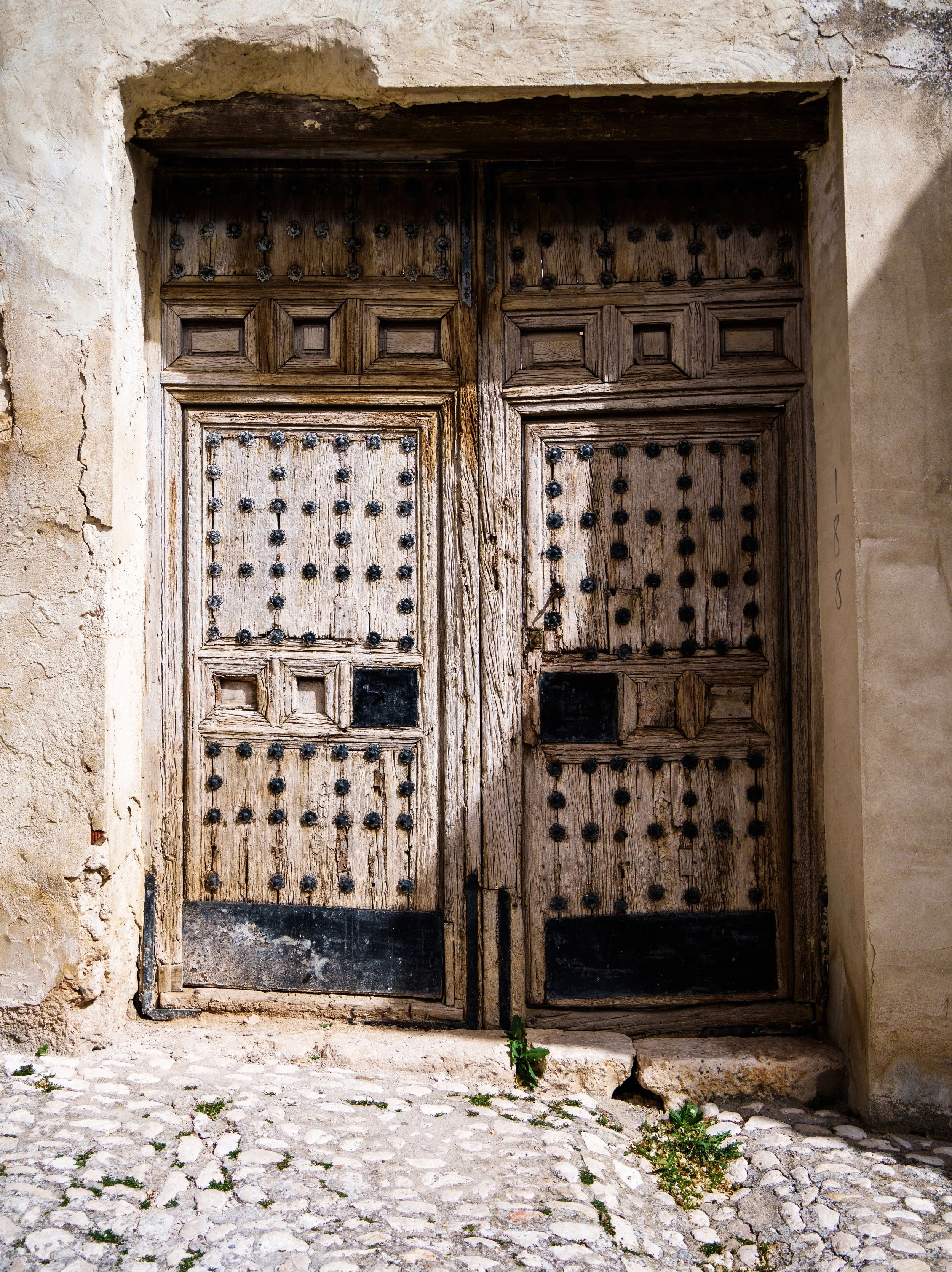 Old wooden double door with iron studs and a weathered, cracked stone wall surrounding it, set in an arched opening. The ground is uneven with stones and small plants growing between them.