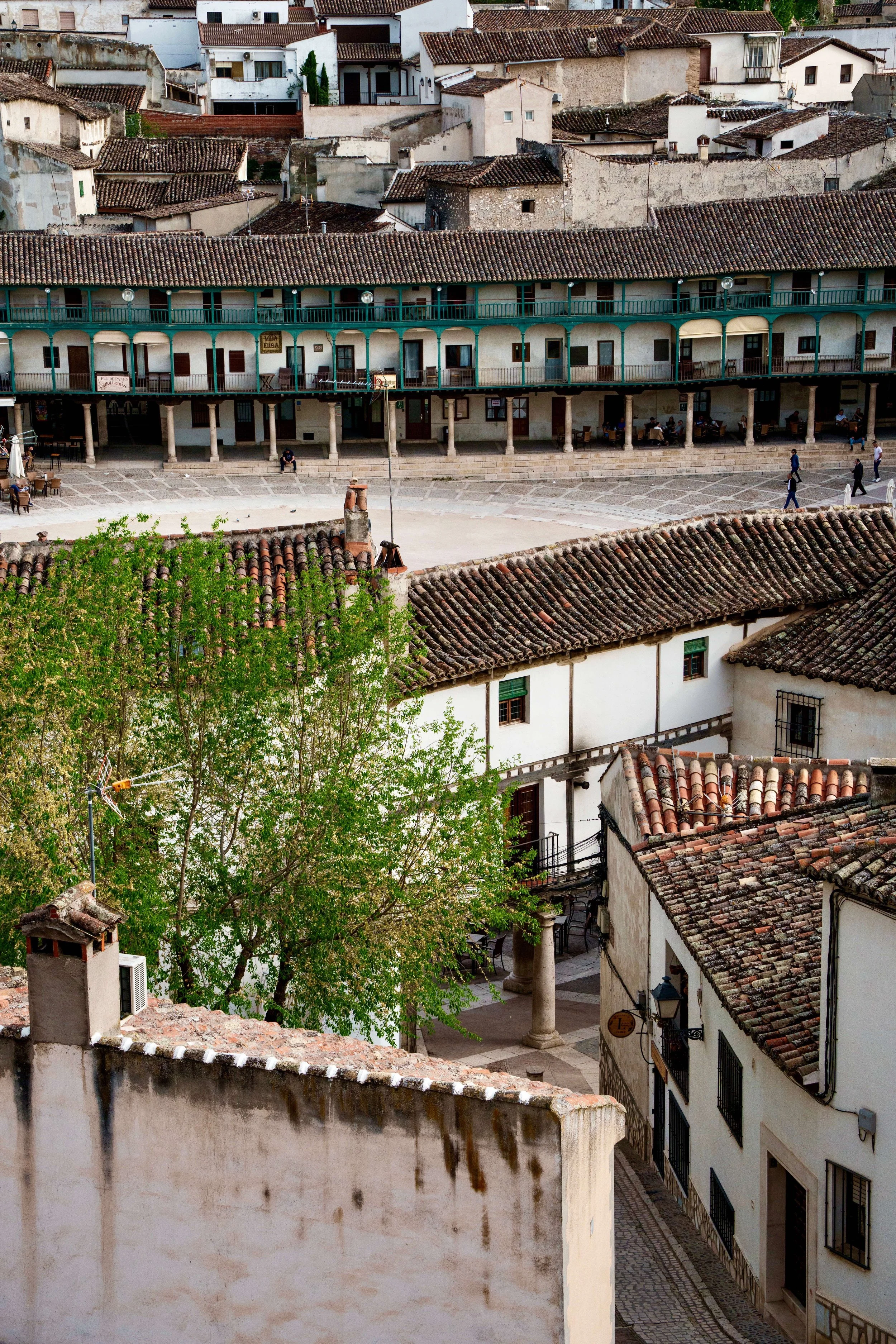 A view of a historic town square with a stone-paved open space, surrounded by white buildings with tiled rooftops. The foreground shows a wall and a cobblestone street, while the background features a row of apartment balconies with green railings an