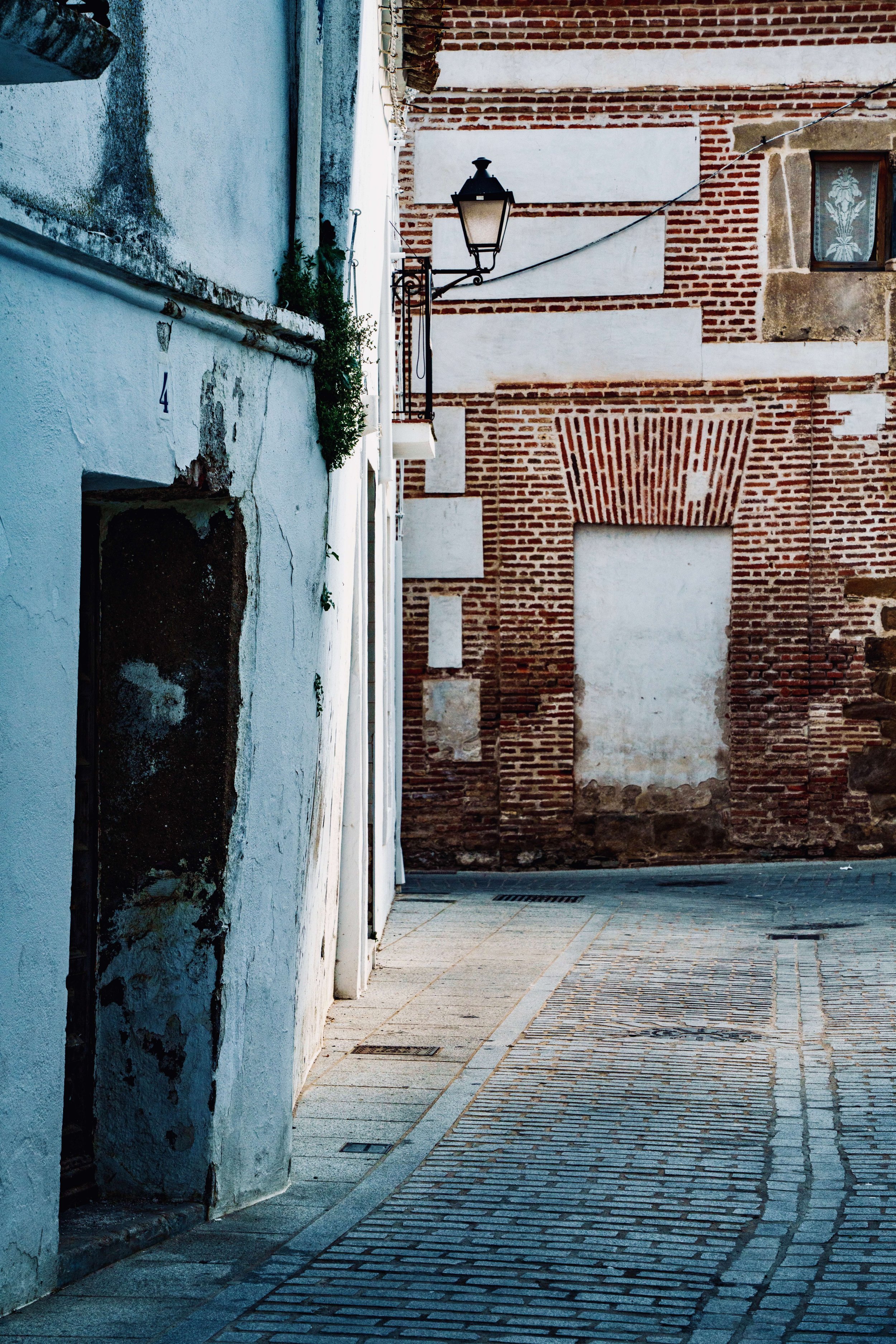 A narrow cobblestone street bordered by white and exposed brick walls, with a black street lamp and a window with lace curtains.