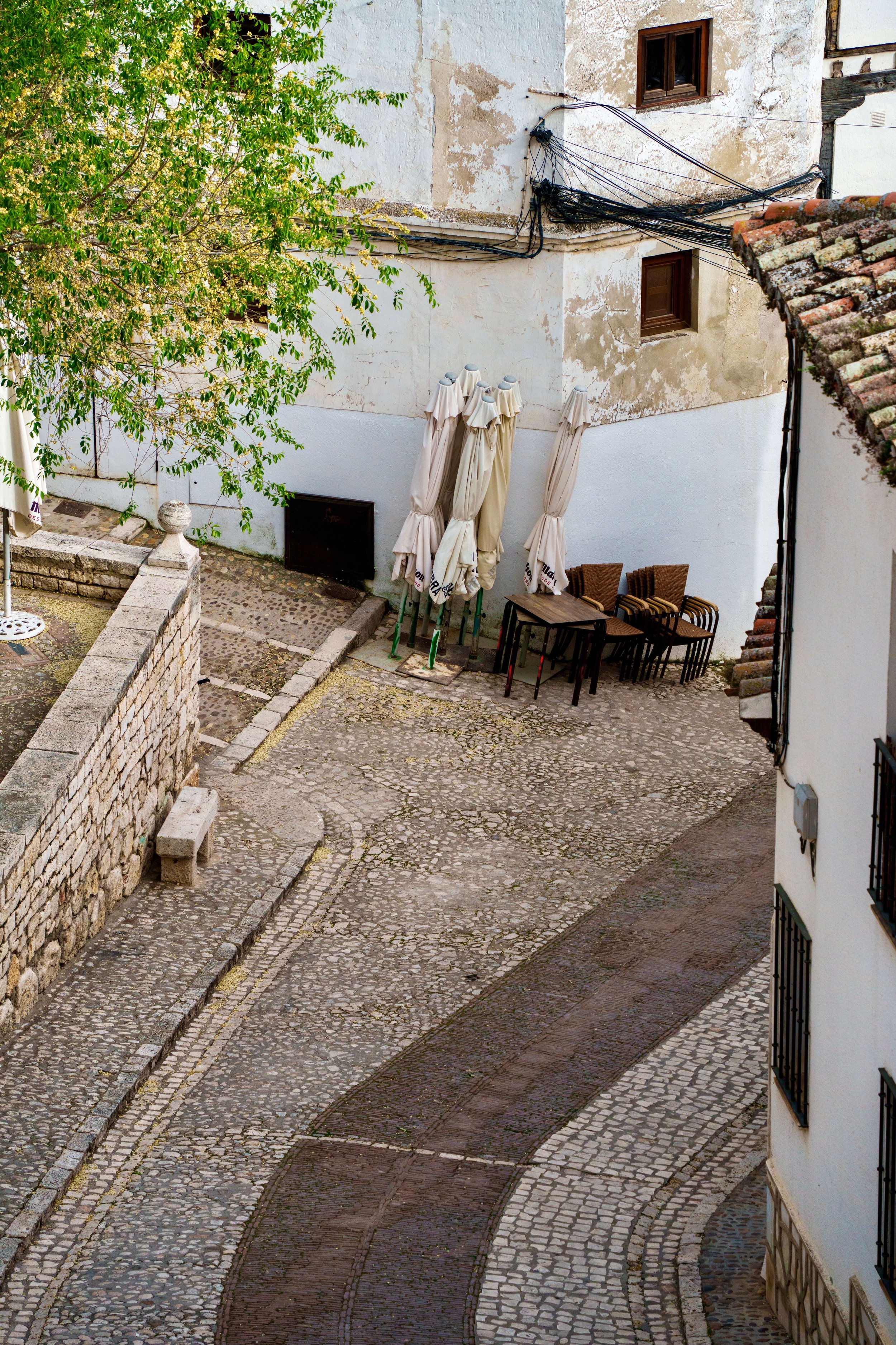 An aerial view of a cobblestone street in a European town with a white building, outdoor tables, umbrellas, greenery, and the roof of the building with vine-covered tiles.