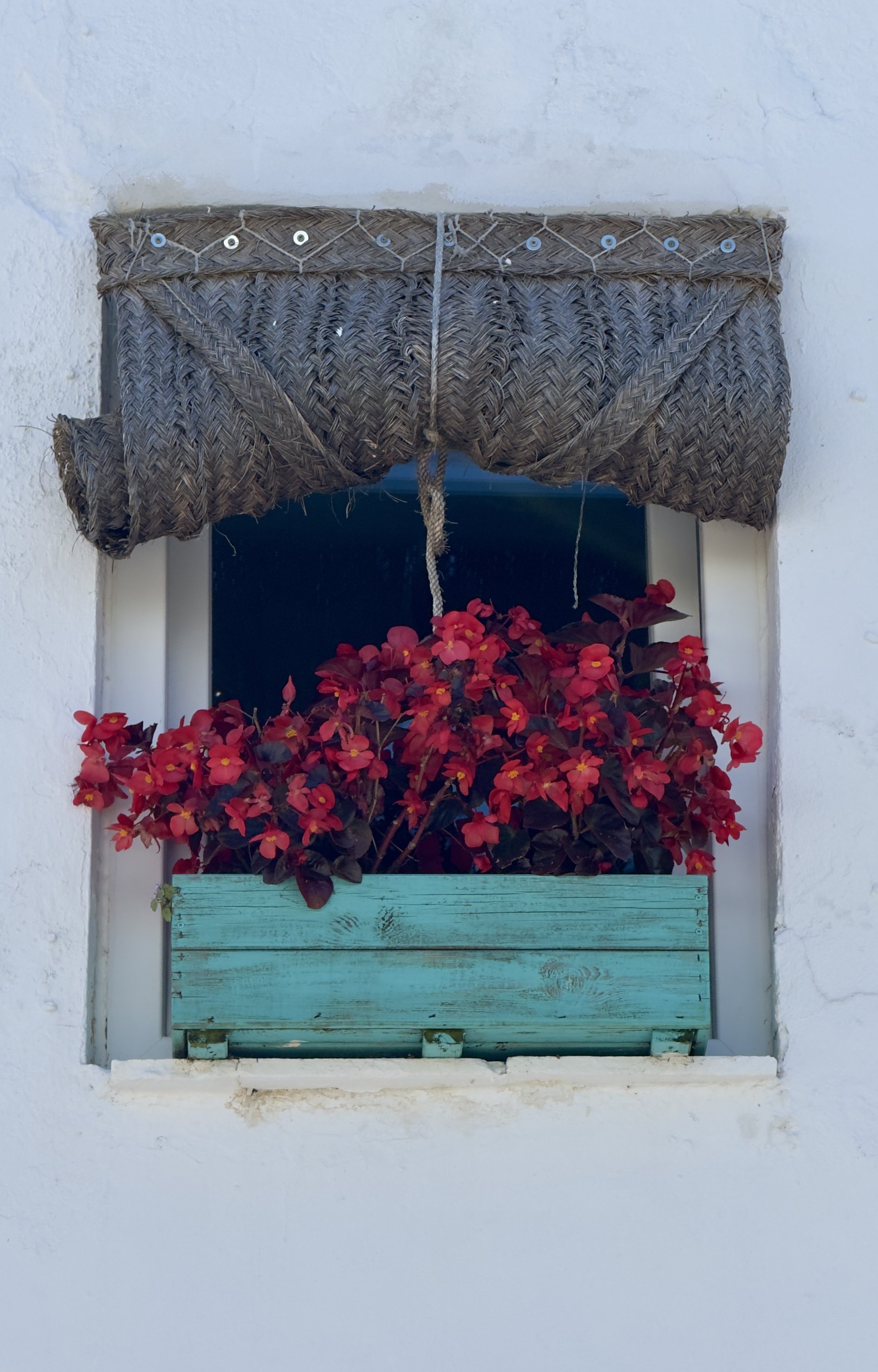 Window with a braided straw shade, a flower box with red flowers, and a turquoise wooden panel beneath the window.