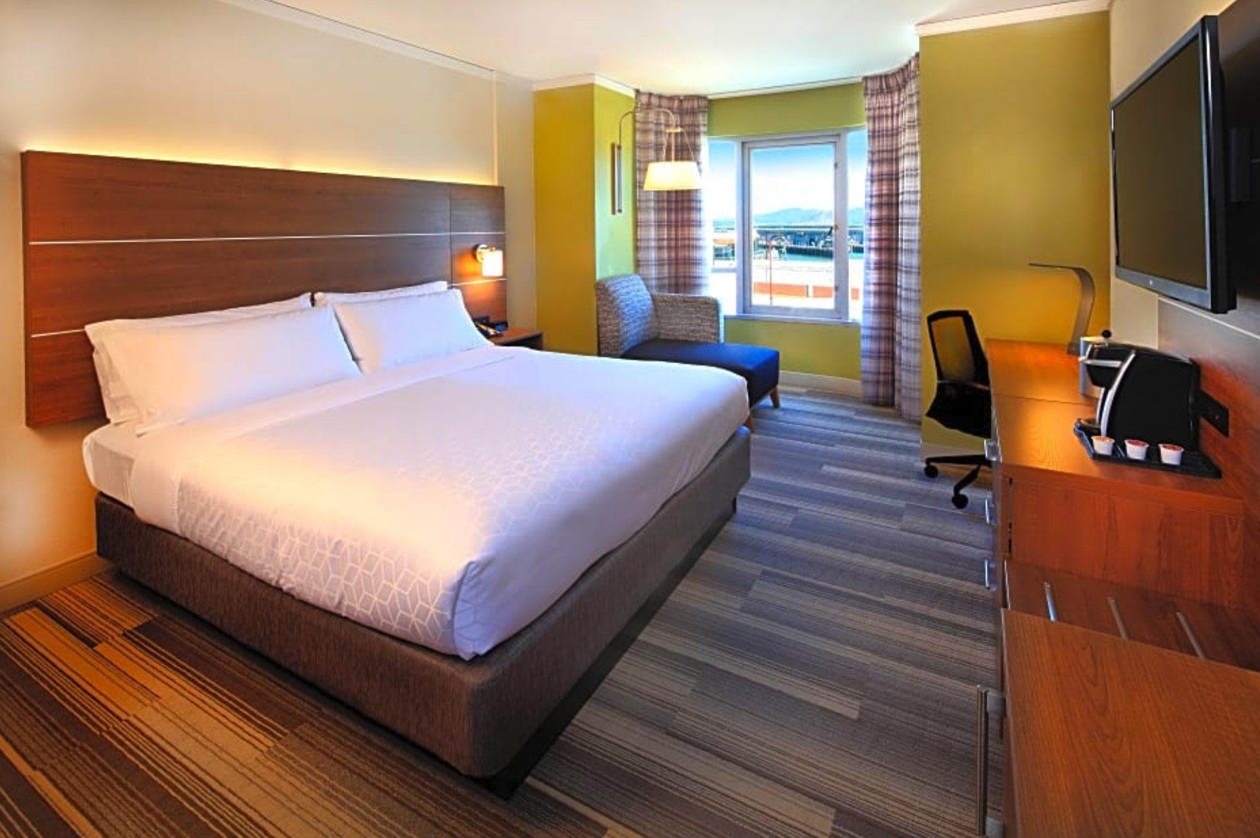 Hotel room with a large bed, window with city view, TV mounted on the wall, and a desk with a coffee maker.
