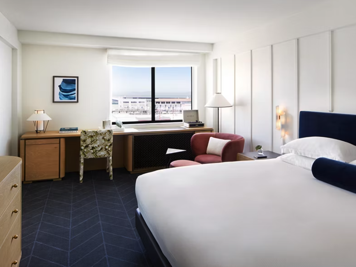 Modern hotel room with a large bed, a pink armchair, a desk with a chair, and a window with a view of a building and the sky.