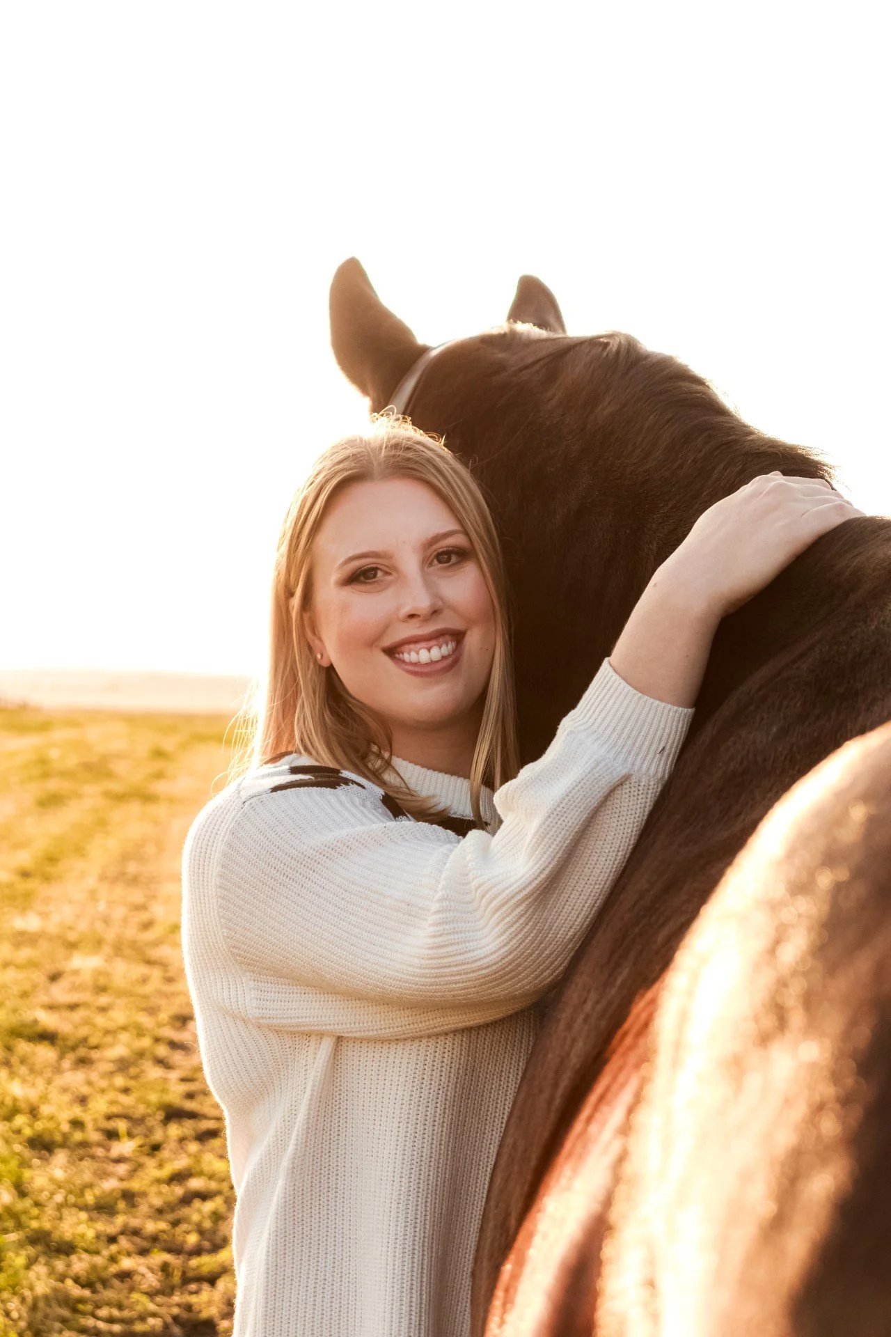 A smiling woman hugging a brown horse outdoors during sunset, in a field with golden grass.