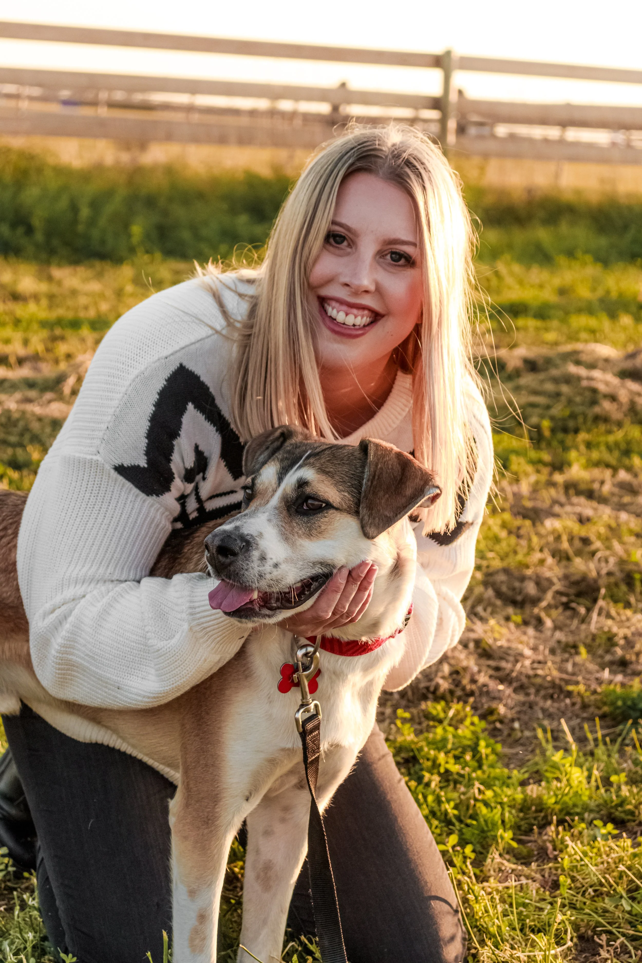 Young woman smiling and holding a dog outdoors during sunset, with a wooden fence and grassy field in the background.