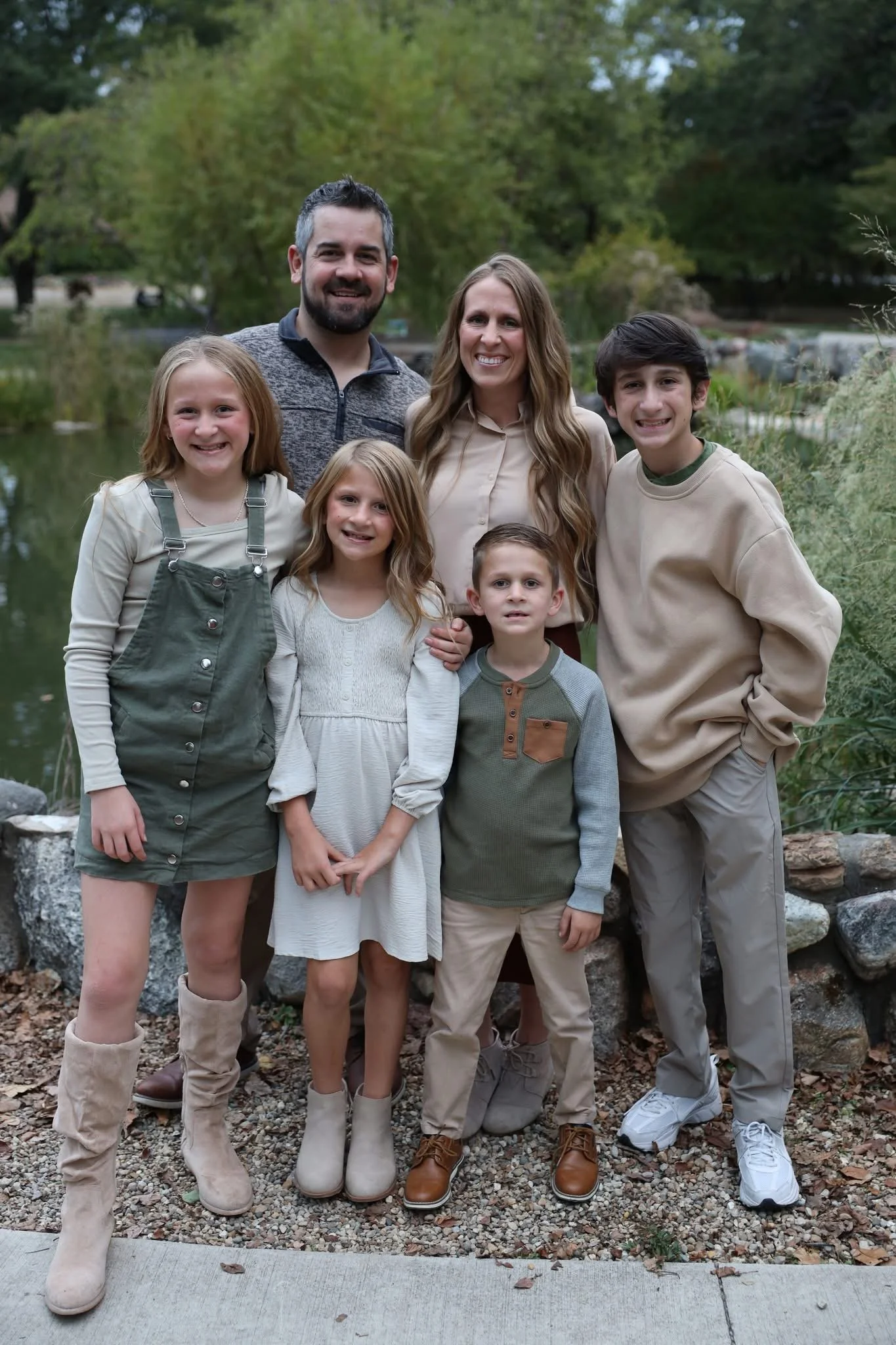 A family of seven people, including a man, woman, and four children, standing outdoors on a rocky and leafy area near a body of water with green trees in the background.