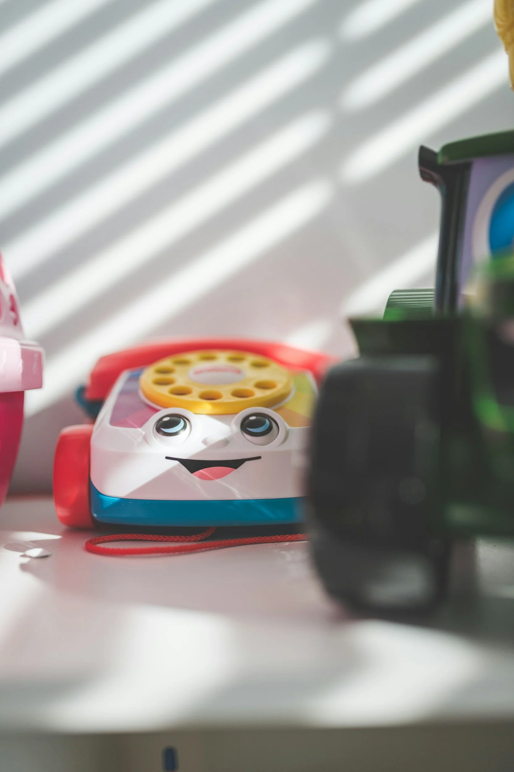 A children's toy phone with a smiling face and big eyes, made of colorful plastic, sitting on a surface with a blurred background including other toys and shadows.