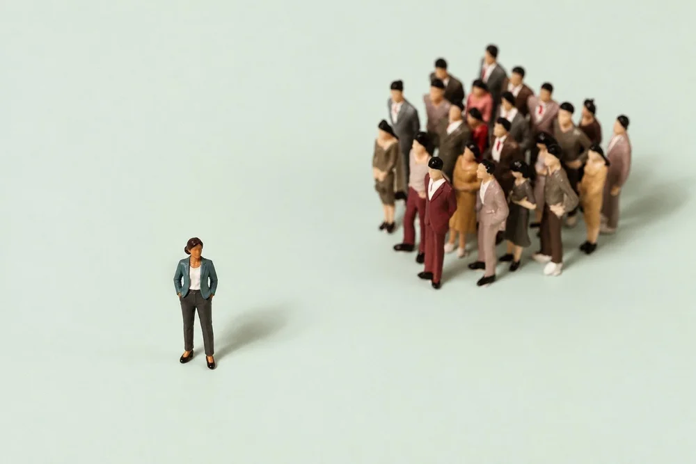 A single miniature figure of a woman of color in a teal blazer standing apart from a large group of figures on a soft sage background