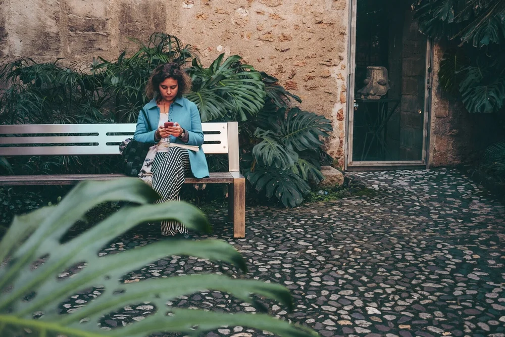 A woman sitting alone on a garden bench, looking down at her phone