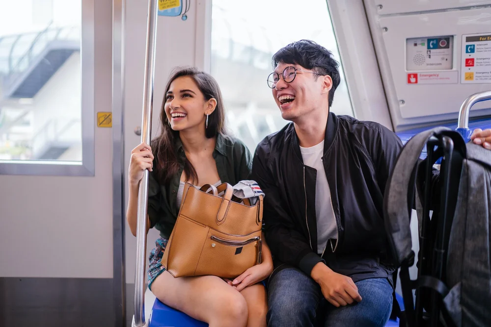 Two friends laughing together on a train, one of them holding her bag on her lap