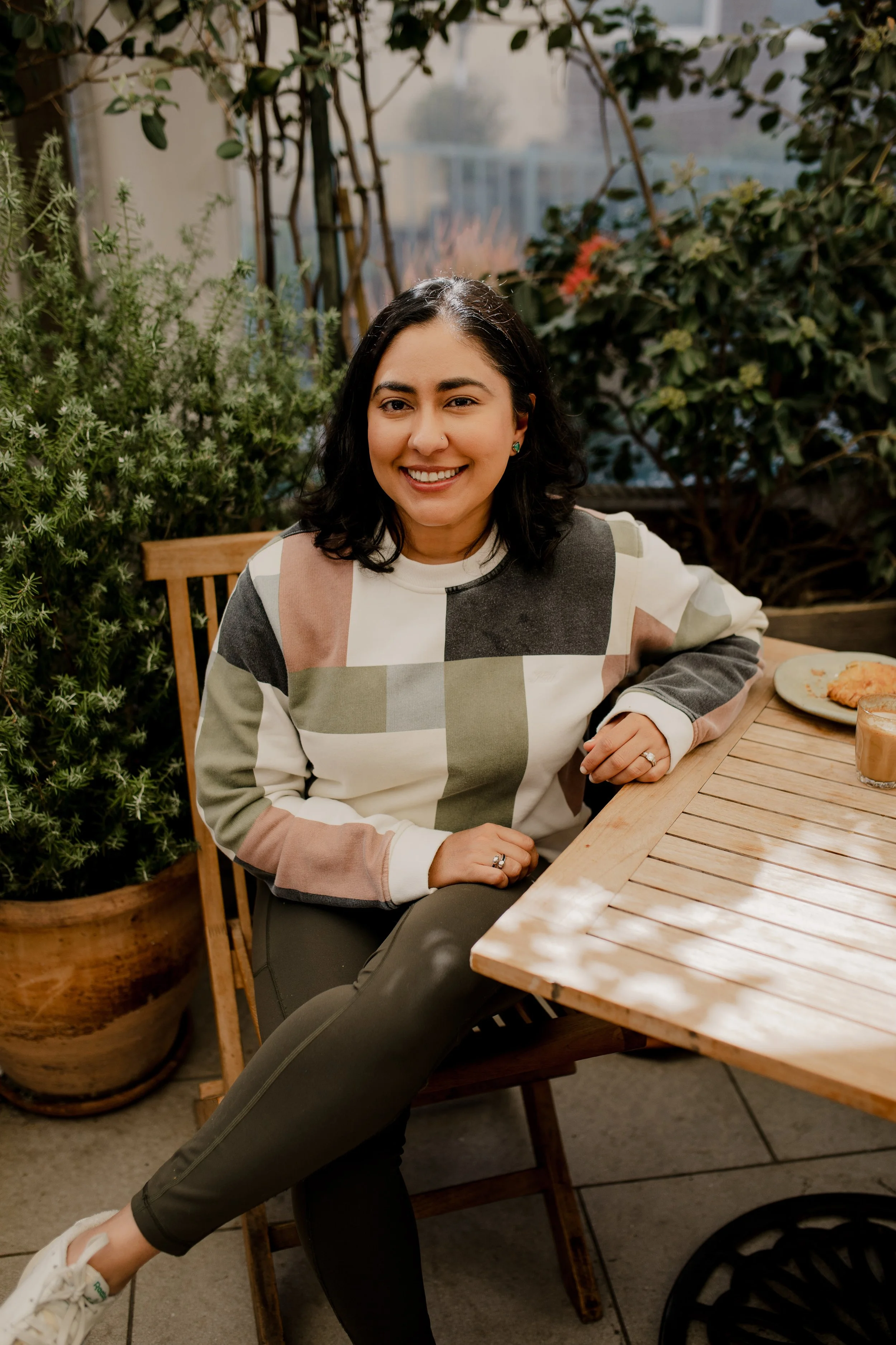A woman with black hair, wearing a patterned sweater, sitting at an outdoor wooden table, smiling, with greenery in the background.