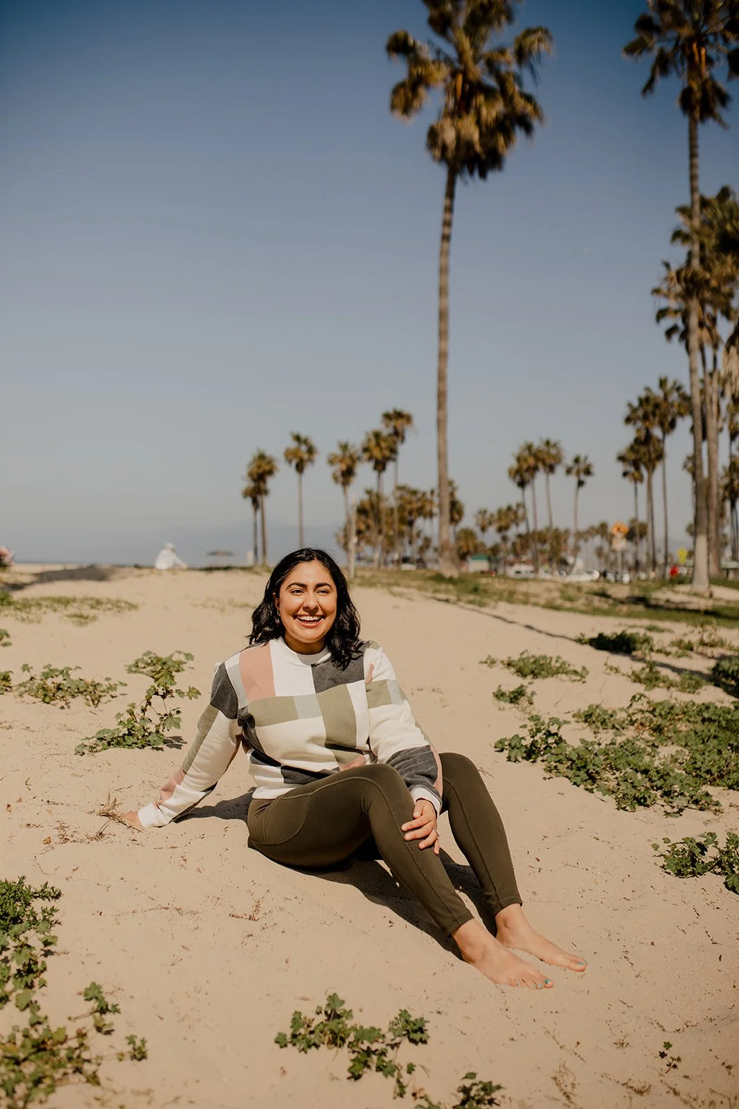 A woman sitting on the sandy beach, smiling, with palm trees in the background and clear blue sky.