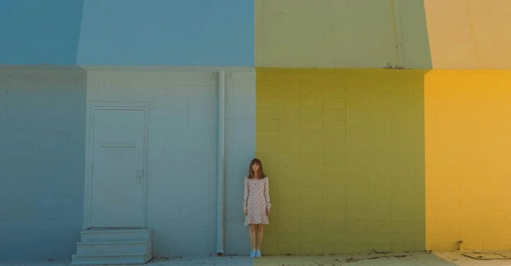 A woman standing alone against a tall wall painted in blue, green, and yellow blocks