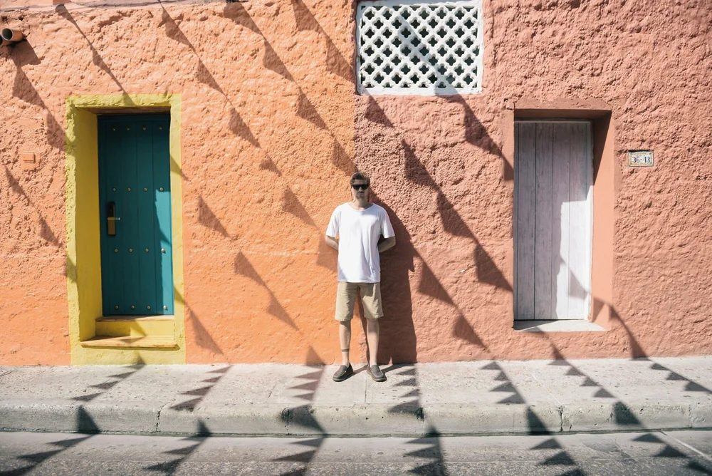 A man in sunglasses and a white T-shirt standing against a warm terracotta wall in strong sunlight