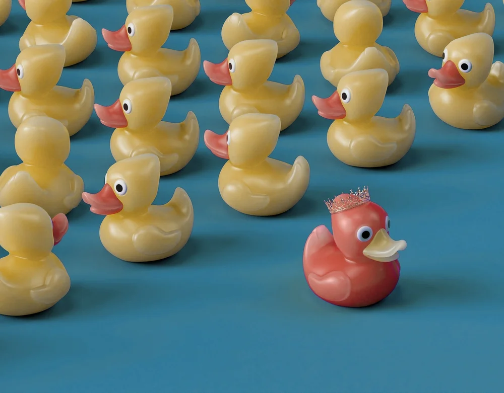 Rows of yellow rubber ducks on a teal surface, with one coral duck wearing a small gold crown set apart from the rest