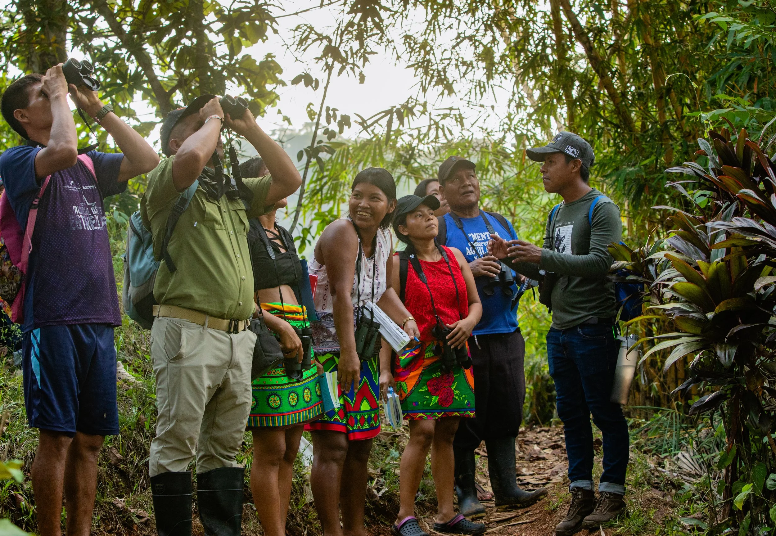 Group of people in a rainforest, some using binoculars and others listening to a guide, dressed in casual outdoor clothing with backpacks and some in traditional patterned skirts.