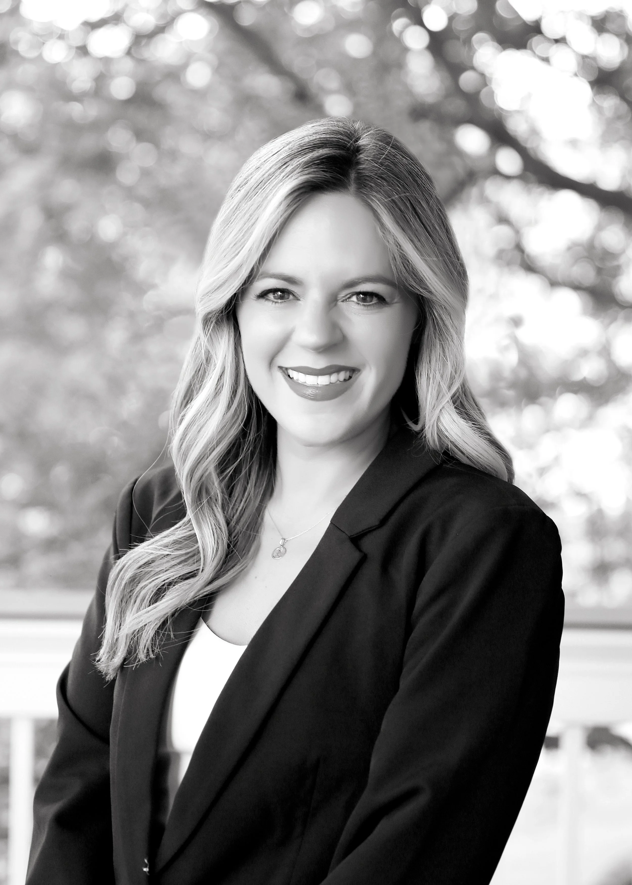 Black and white portrait of a smiling woman with long wavy hair, dressed in a blazer and white top, outdoors with a blurred natural background.