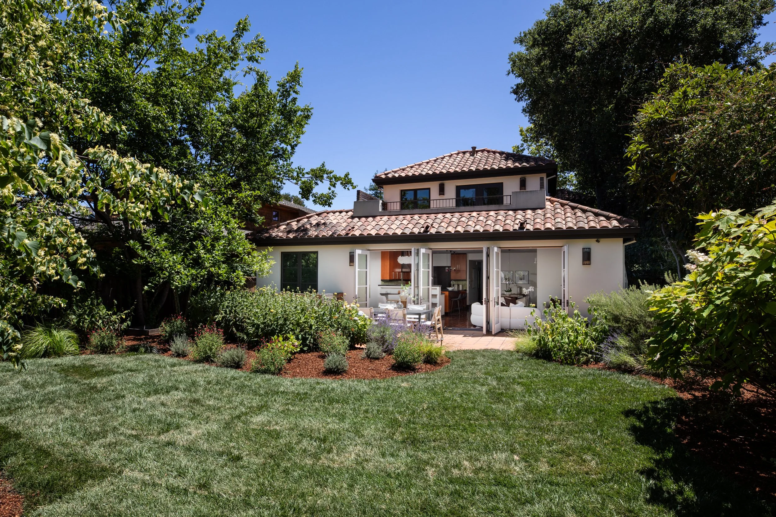 A backyard garden with a well-maintained green lawn, flowering bushes, and mature trees surrounding a modern two-story house with a tiled roof and open sliding glass doors revealing an indoor living space.