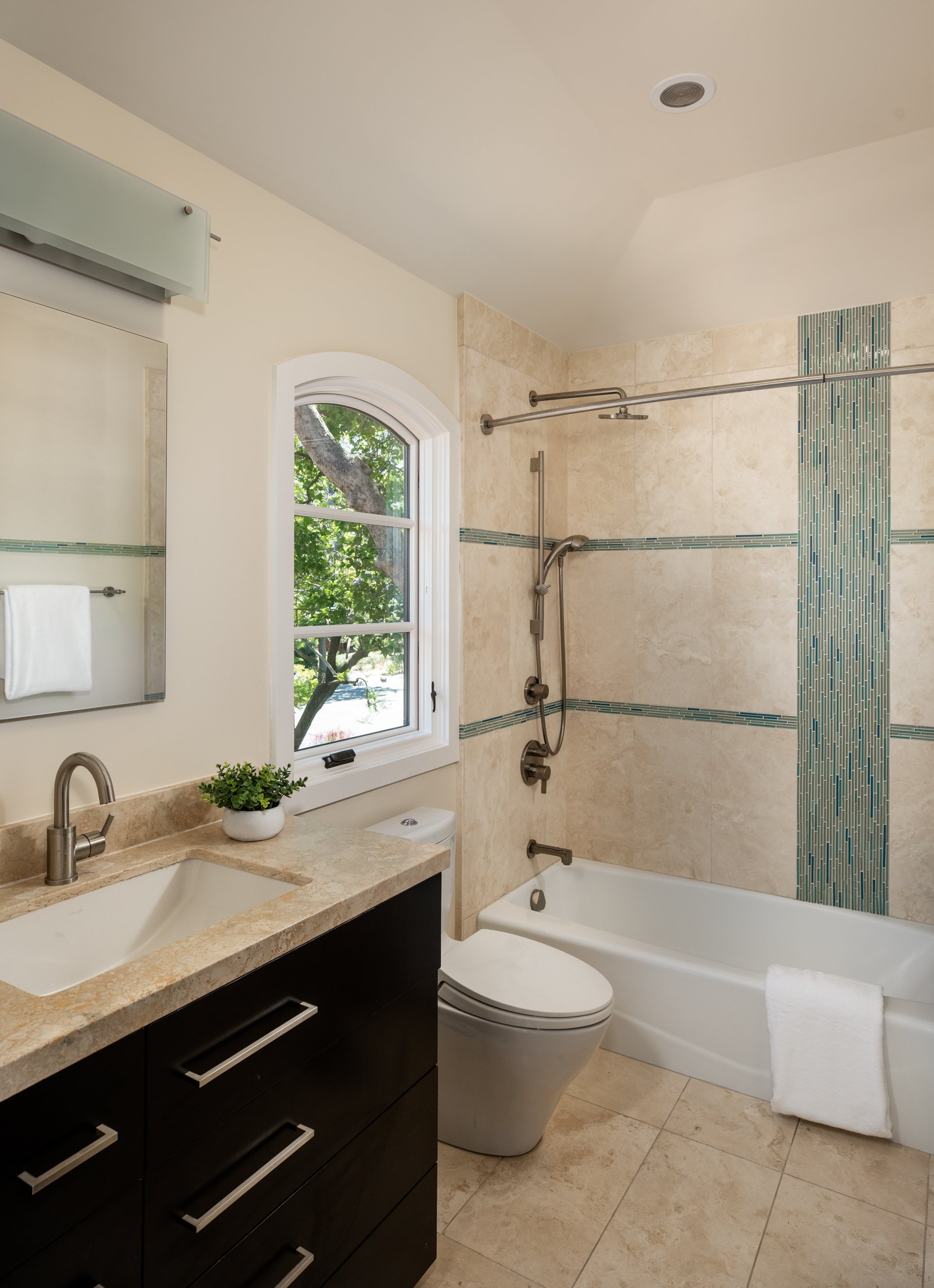 Modern bathroom with beige tile walls and floor, a white bathtub, a black vanity with a beige marble countertop, a small plant, a mirror, a window with greenery outside, and a shower area with a rod and patterned tiles.