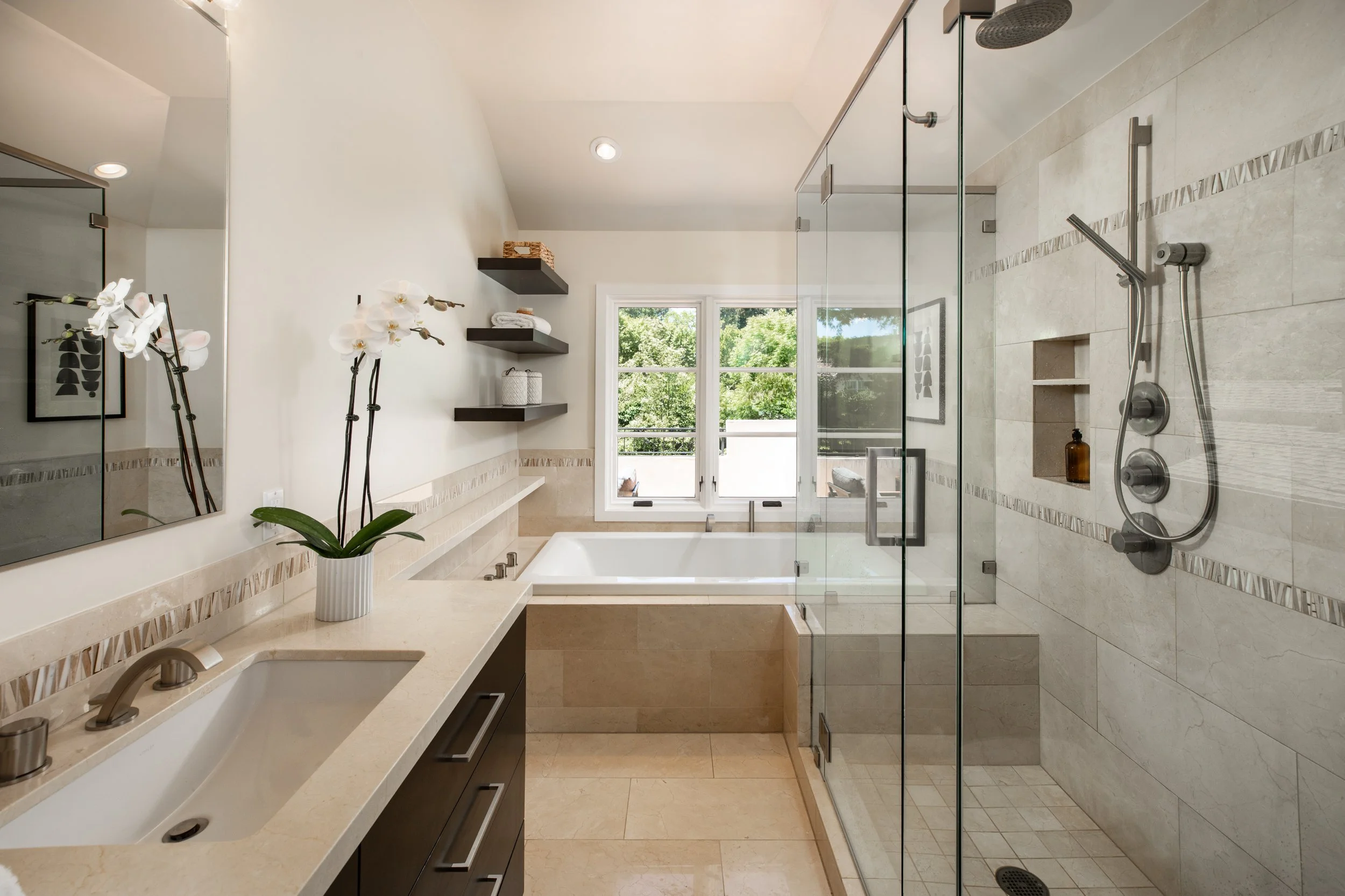 Modern bathroom with a walk-in shower, bathtub, white walls, beige tiled floor, and large window overlooking greenery, decorated with floating black shelves and a potted orchid.