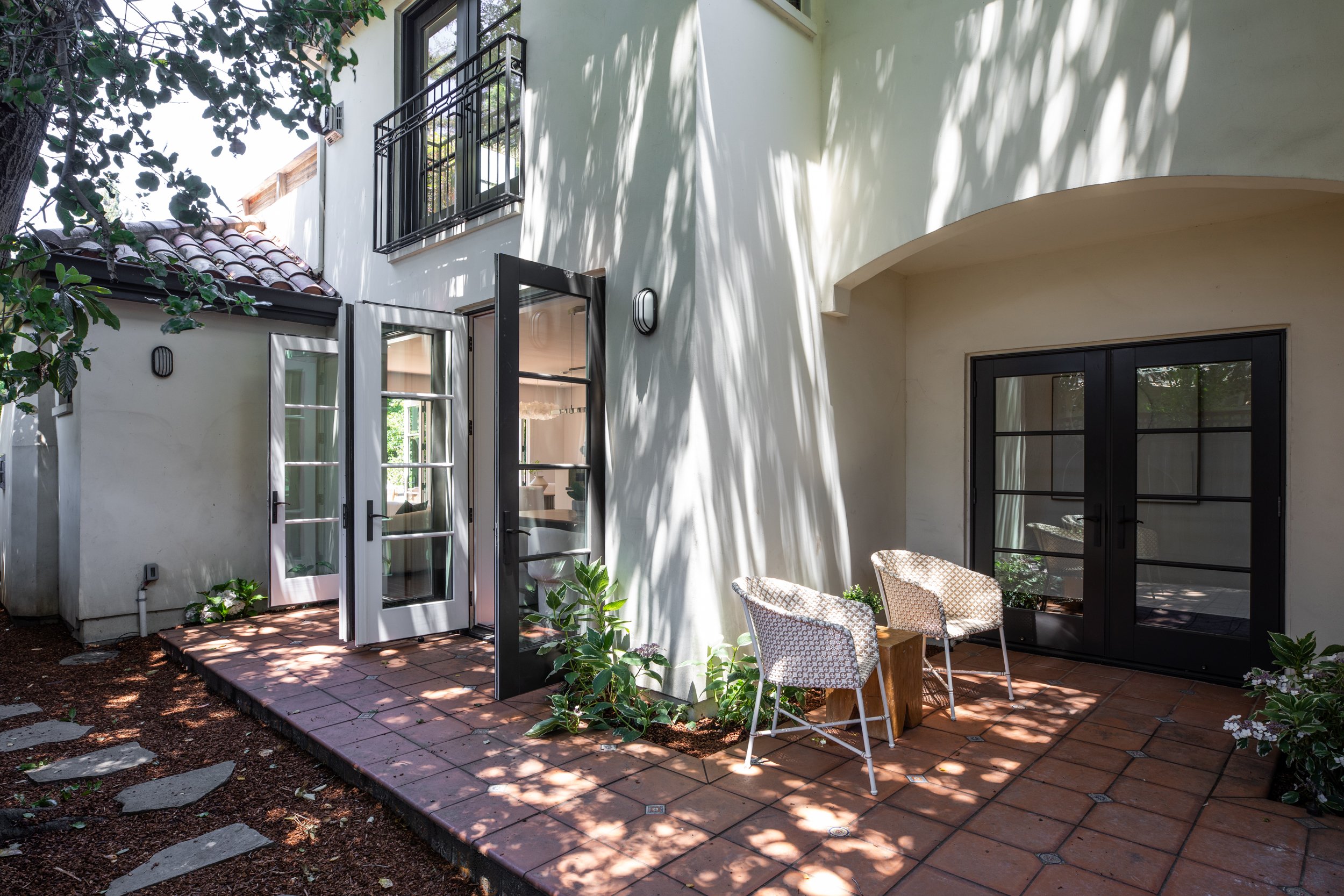 A shaded patio area with a brick floor and two white chairs with patterned cushions, adjacent to a white house with black-framed windows and an open door. There are plants and trees providing shade.