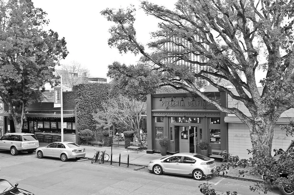 Street view with parked cars, bicycles, trees, and a restaurant named Pizzeria Delfina on the right side of the image.