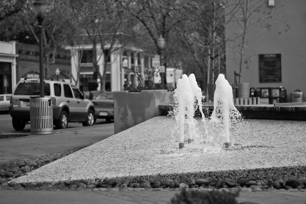 A small tabletop fountain with two water jets in an urban park setting, with parked cars, trees, and buildings in the background.