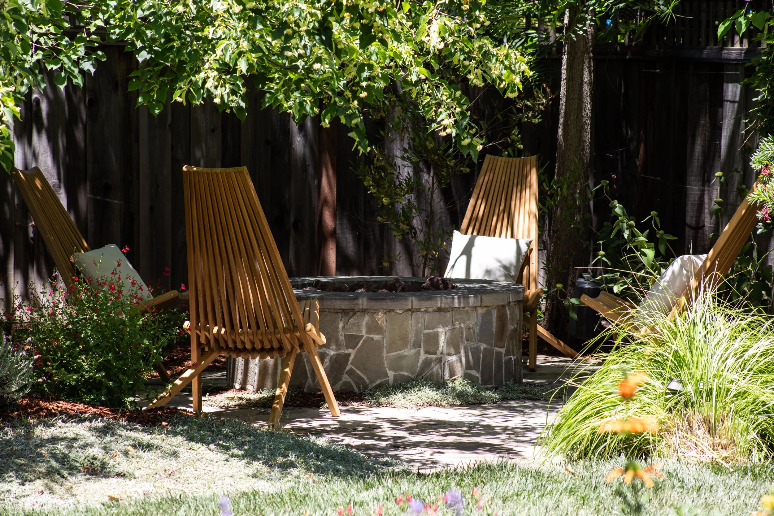 A backyard garden with a stone fire pit surrounded by four wooden chairs. The area is shaded with lush green leaves and plants, with a wooden fence in the background.