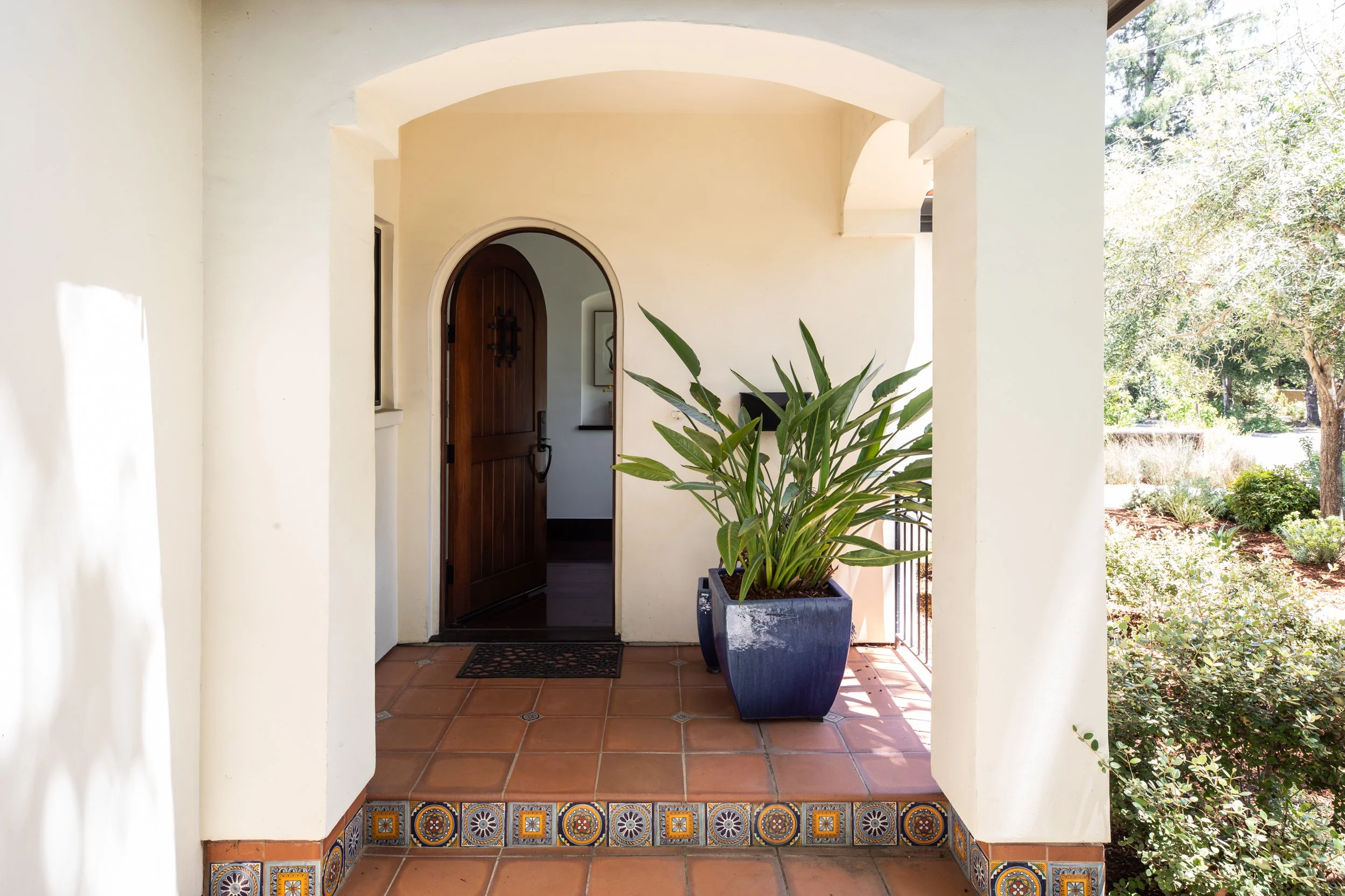 Entryway of a house with a wooden door, potted plant, and decorative tile border on the steps, overlooking a garden with trees and shrubs.