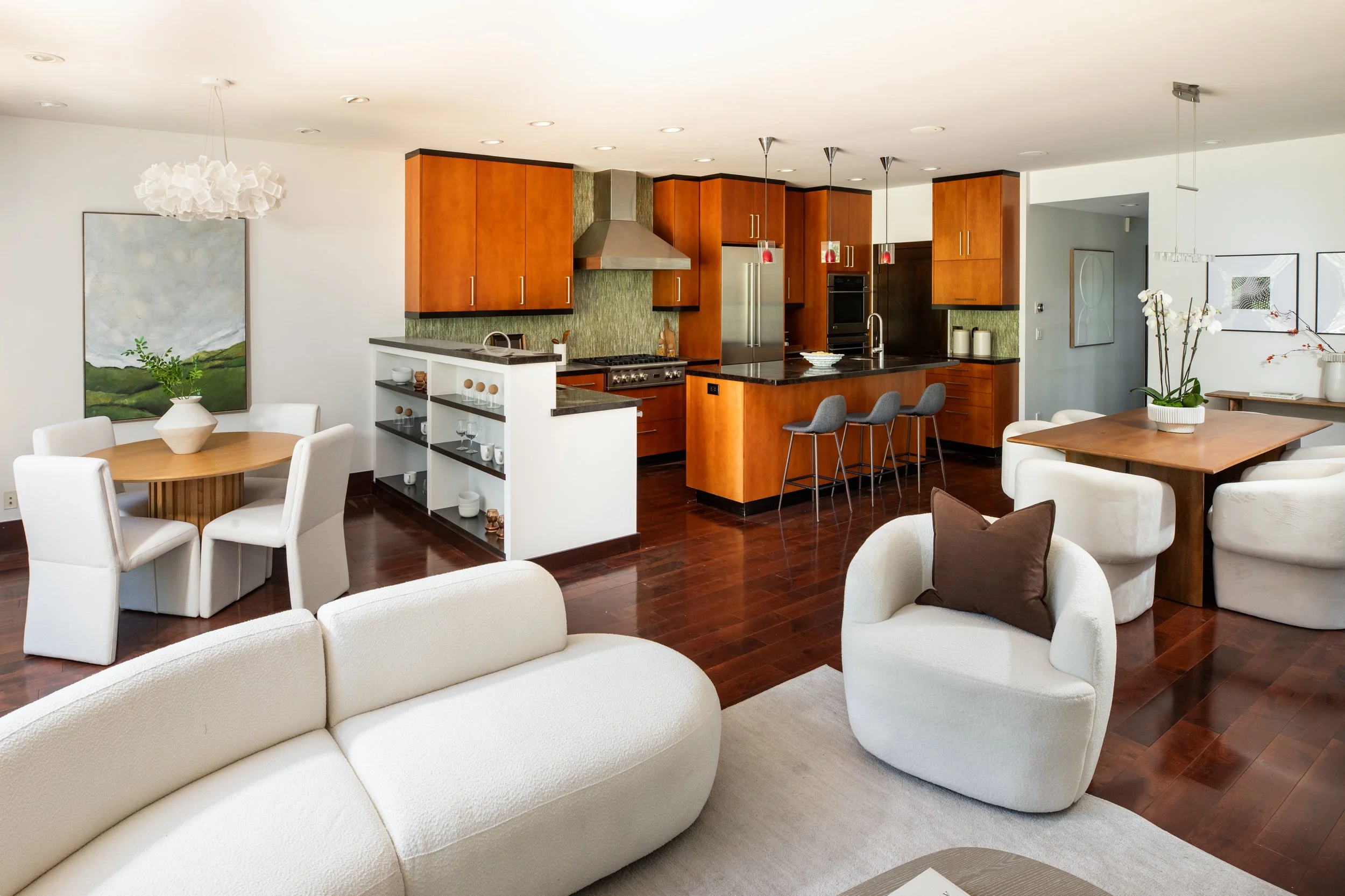 Open-concept living room and kitchen with white curved sectional sofa, dining table with white chairs, wooden kitchen cabinets, stainless steel appliances, green textured backsplash, and decorative wall art.